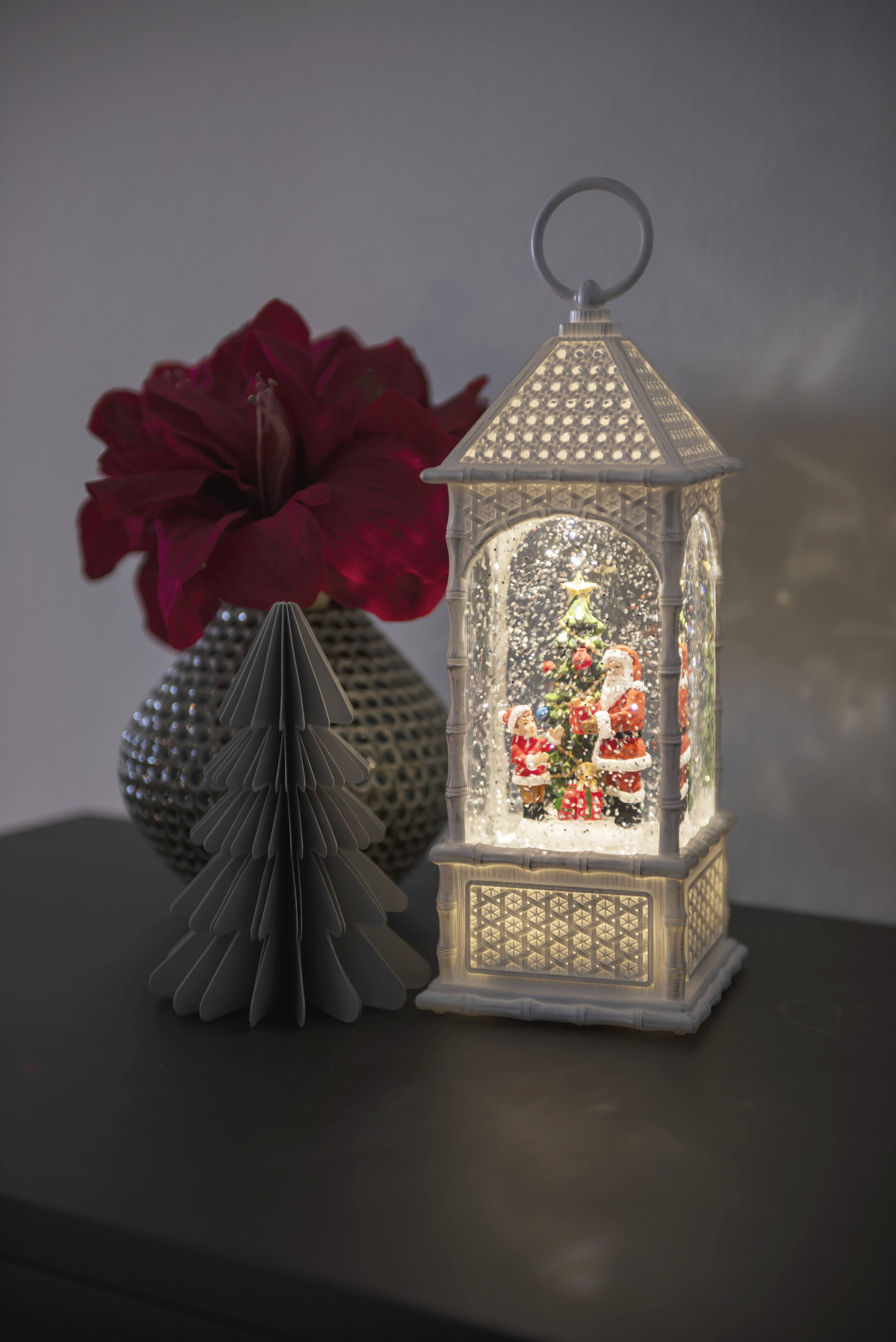 Ein leuchtender Weihnachtslaterne mit Schneegestöber und Weihnachtsmann-Figur steht auf einem Tisch neben einer roten Blume und einer Deko-Tanne.