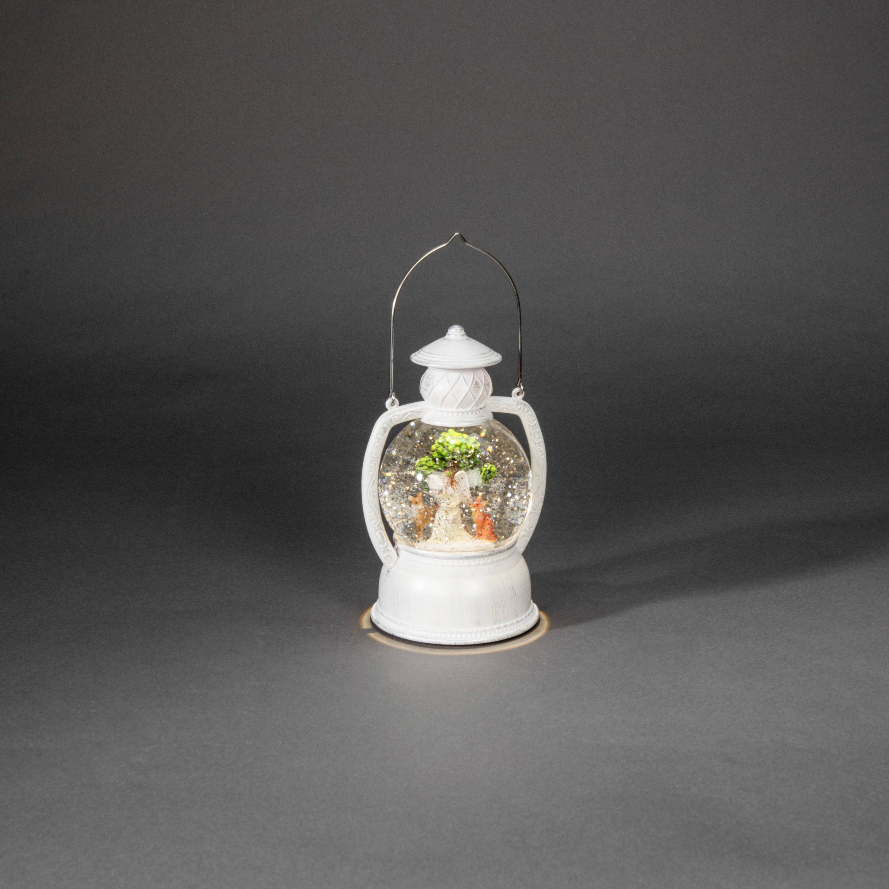 Eine kleine, weiße Laterne mit abgerundetem Griff. Innen sind winzige Ornamente, darunter ein Weihnachtsbaum und Geschenke.