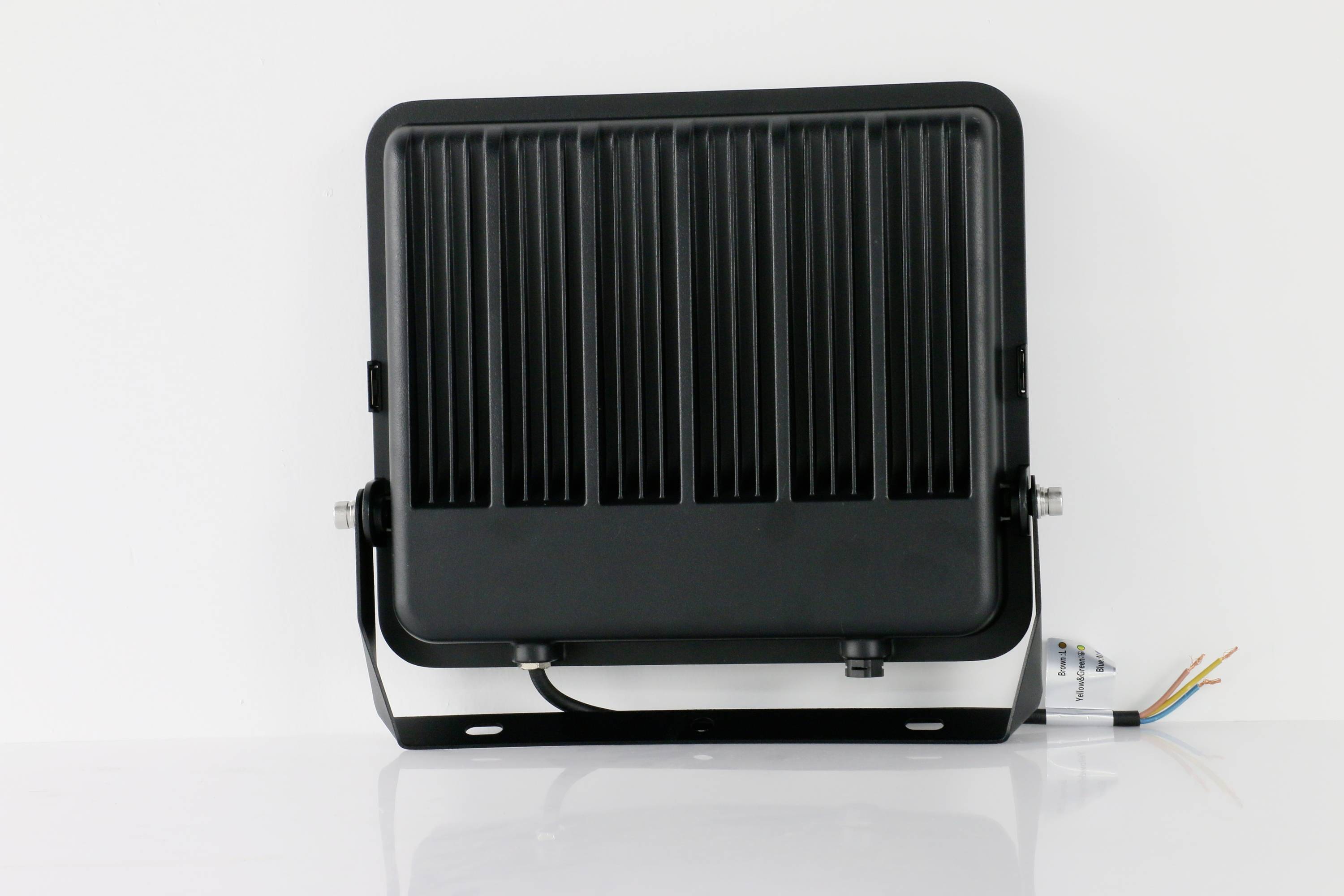 Ein schwarzer LED-Flutlichtstrahler mit geripptem Gehäuse steht auf einer weißen Oberfläche. Rechts sind bunte Kabel sichtbar.