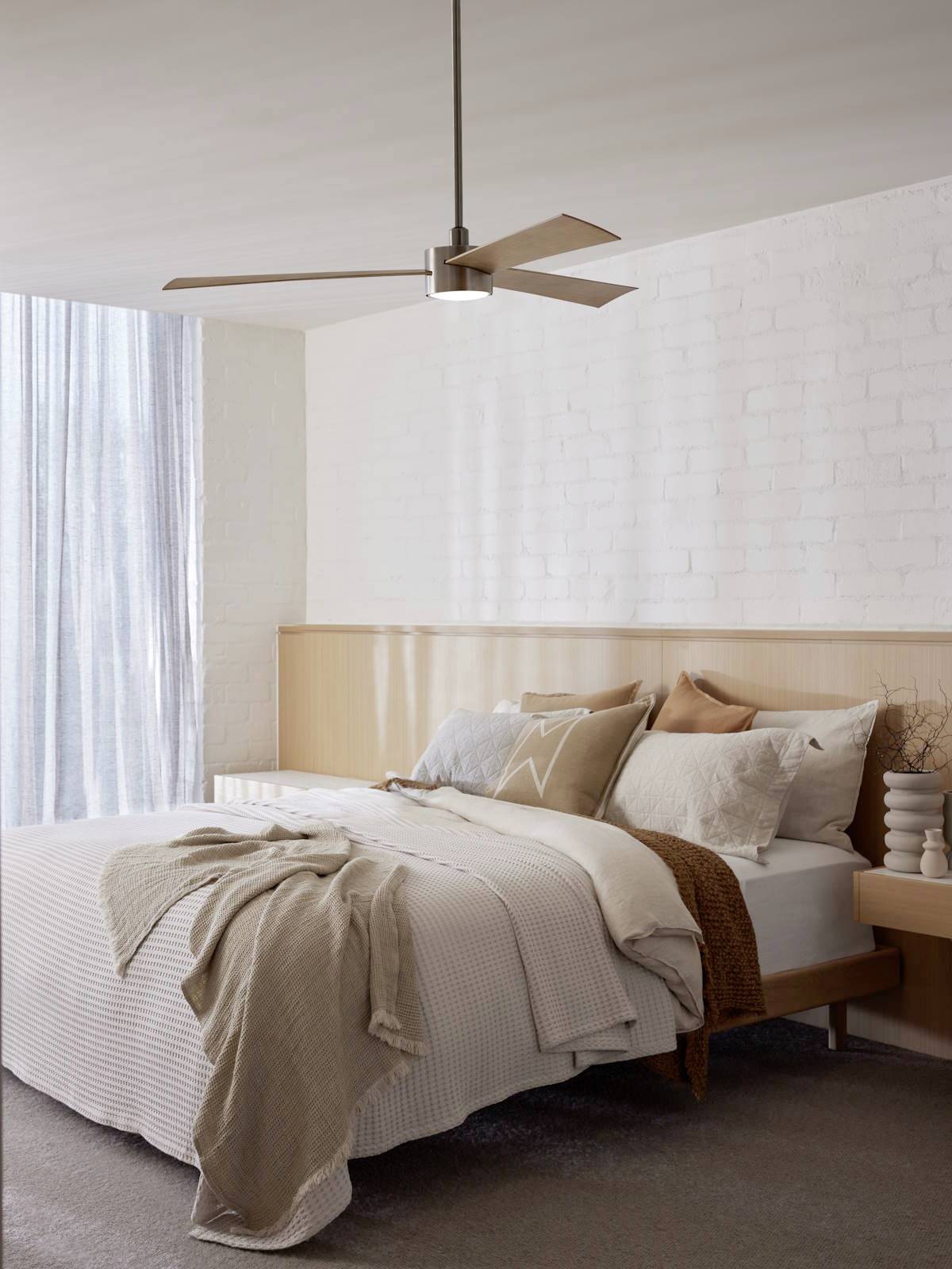 Ein modern eingerichtetes Schlafzimmer mit einem Bett, das mit mehreren Kissen und einer beigen Decke dekoriert ist. Helle Wände und ein Deckenventilator ergänzen das minimalistische Design.