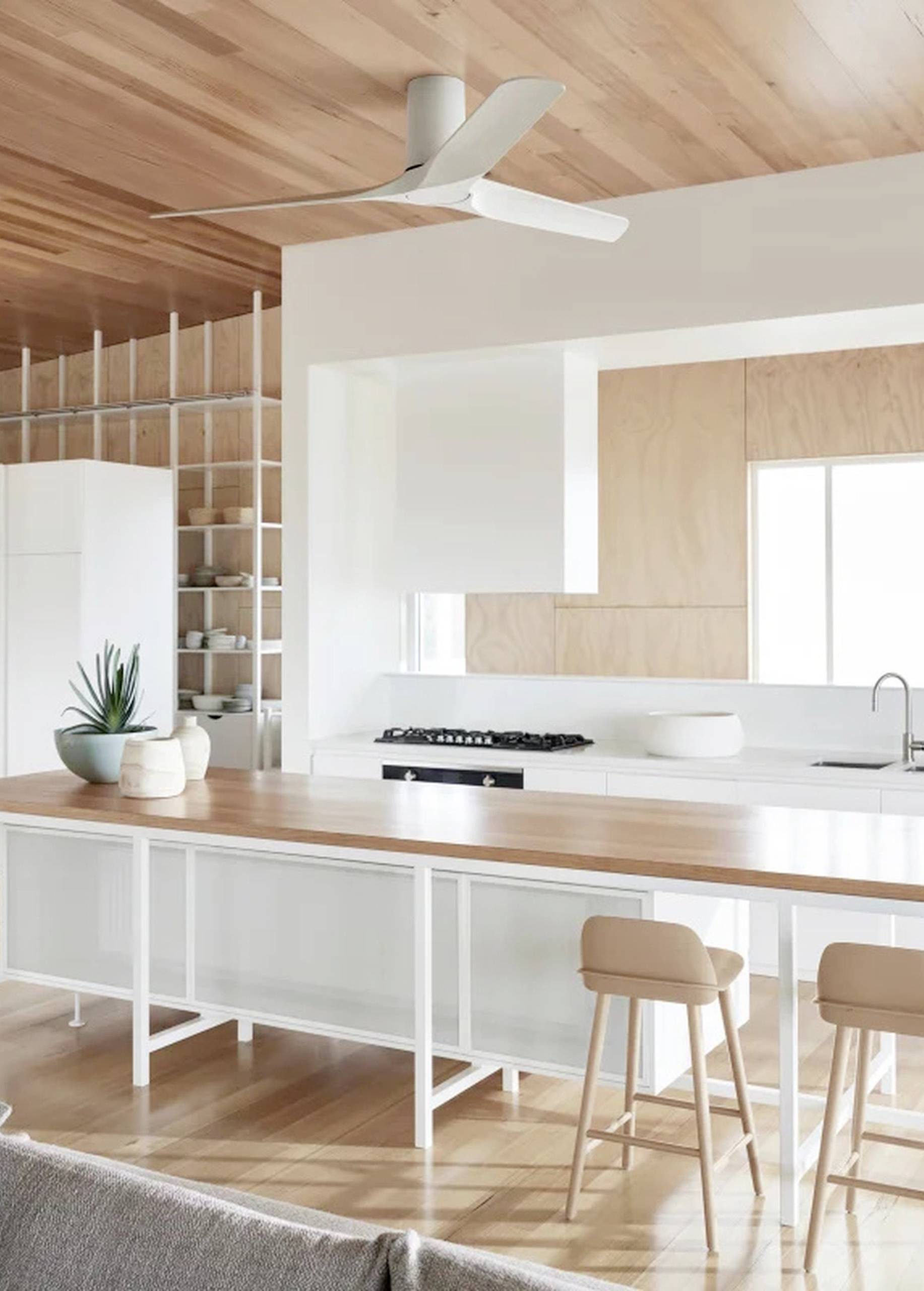 Moderne Küche mit Holzdecke und Insel. Weiße Schränke und Barhocker, Pflanzenakzent und großes Fenster sorgen für helle Atmosphäre.