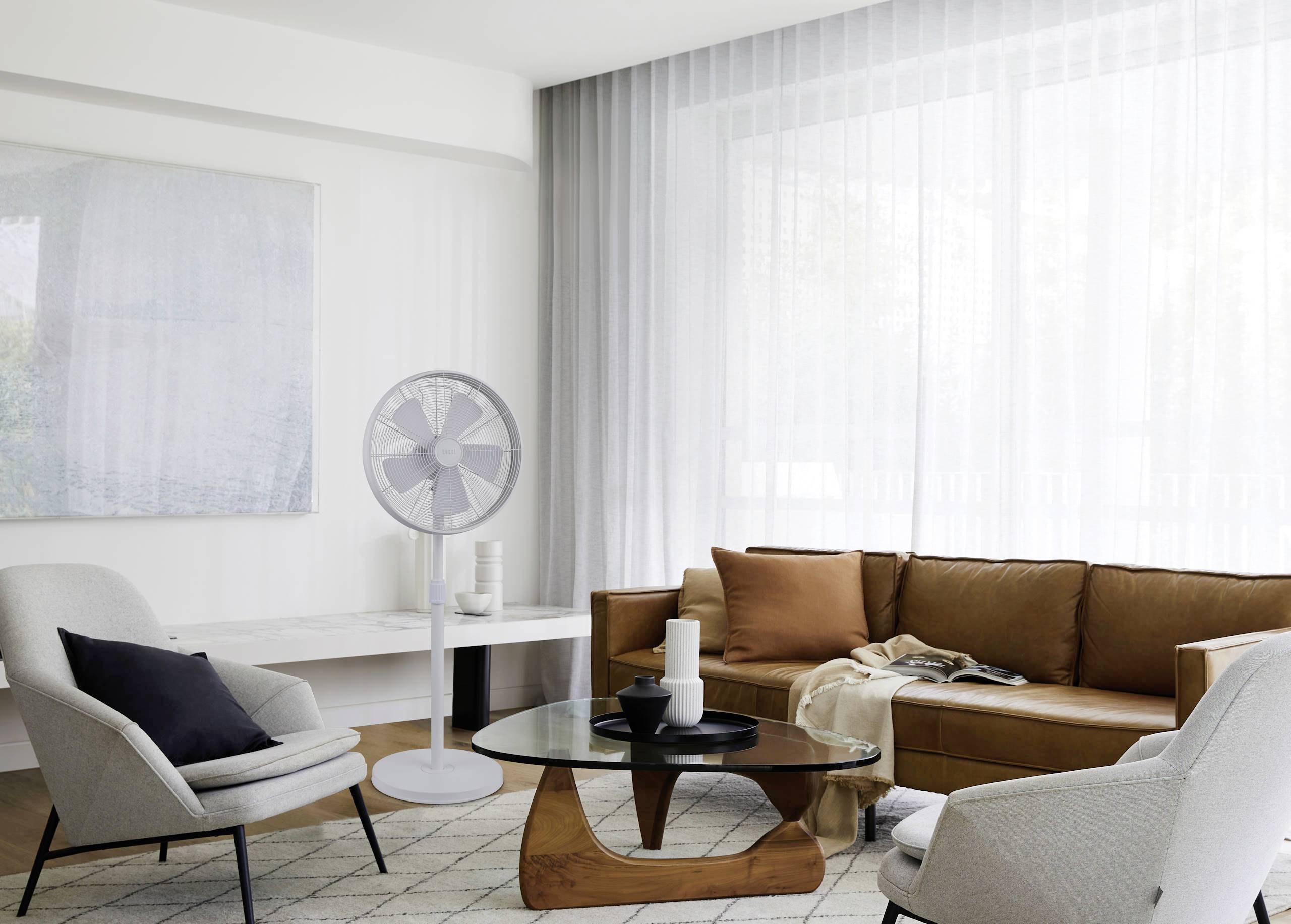 Ein modernes Wohnzimmer mit einem Ledersofa, Sesseln, einem Glastisch und Ventilator. Große Fenster mit weißen Vorhängen lassen Licht herein.