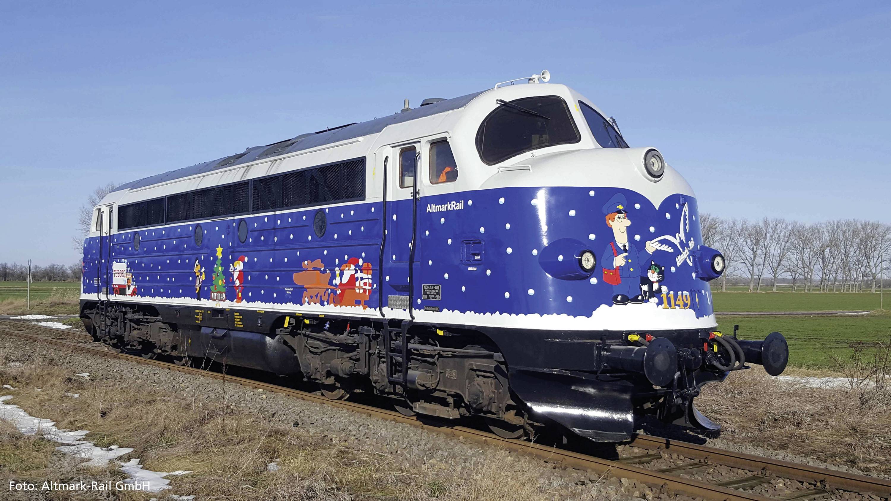 Eine blau-weiße Lokomotive mit winterlichen und weihnachtlichen Motiven fährt auf einer ländlichen Bahnstrecke unter klarem Himmel.