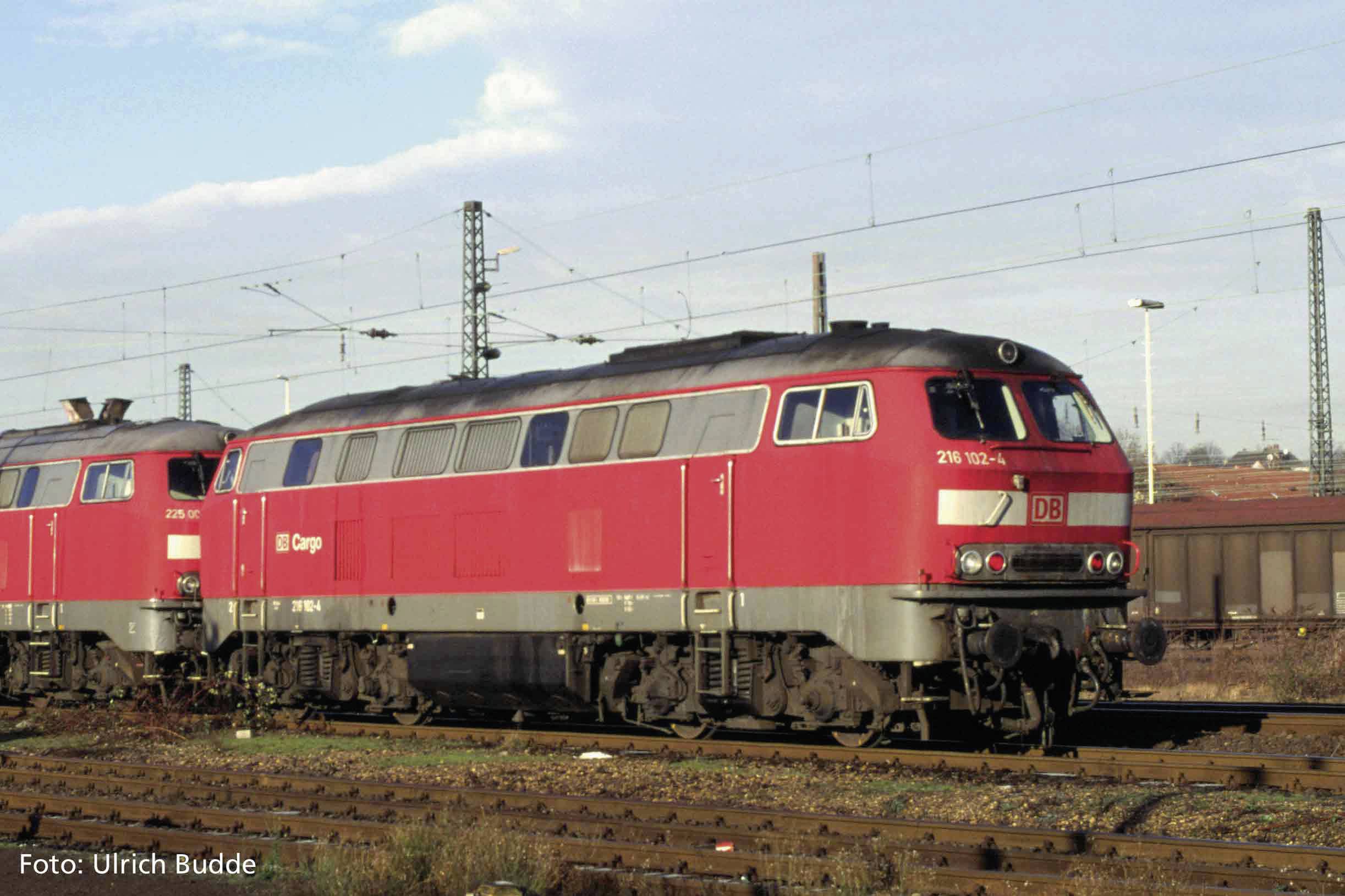Roter Zug der Deutschen Bahn auf einem Bahngleis, mit anderen Zügen im Hintergrund. Es ist ein bewölkter Tag, Stromleitungen sind sichtbar.