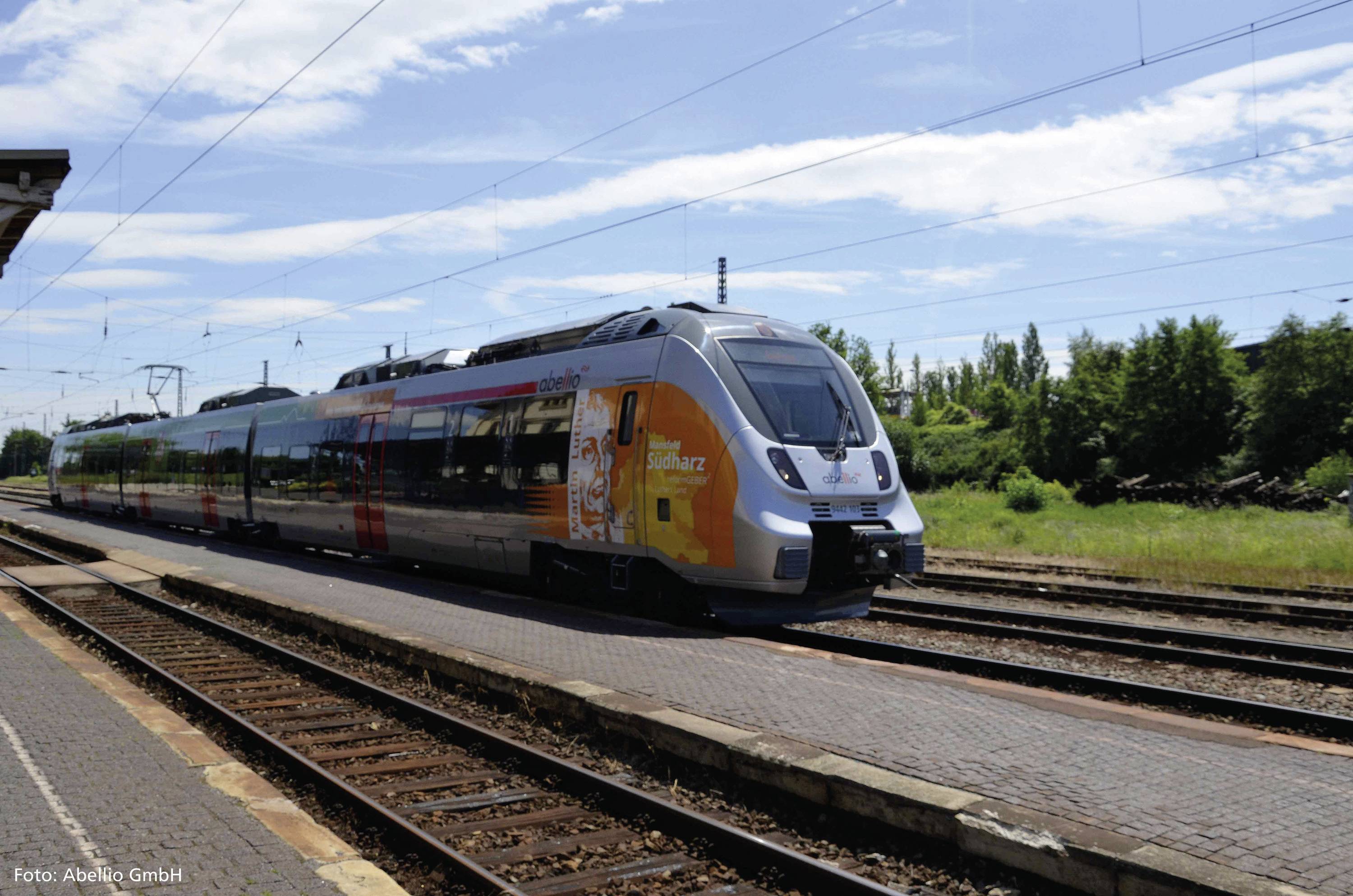 Ein moderner Regionalzug fährt auf einem sonnigen Bahnsteig entlang. Im Hintergrund sind Bäume und blauer Himmel zu sehen.