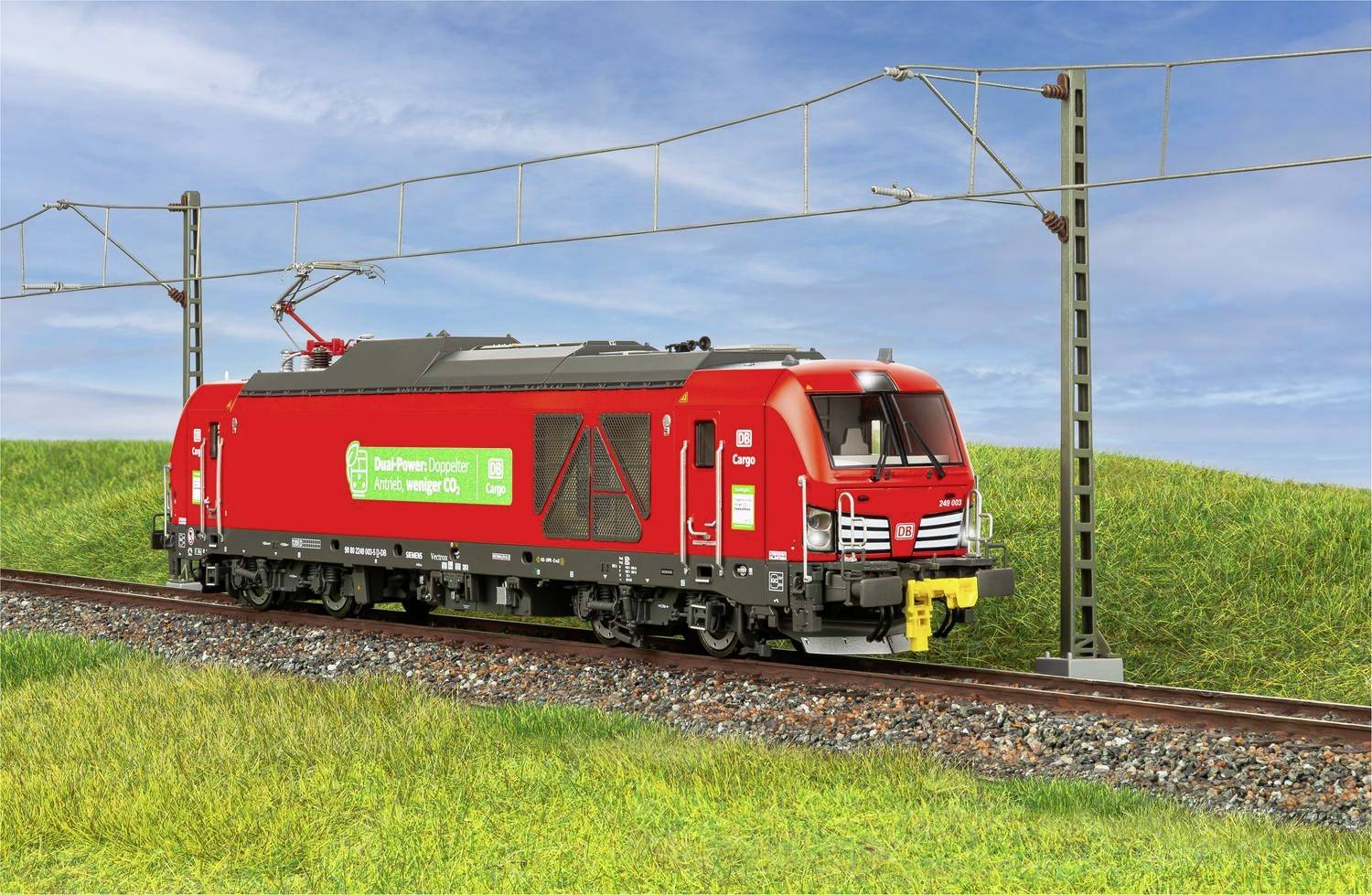 Eine rote Güterzuglokomotive fährt auf Schienen entlang einer grünen Wiese unter bewölktem Himmel. Sie ist von 'DB Cargo' beschriftet.