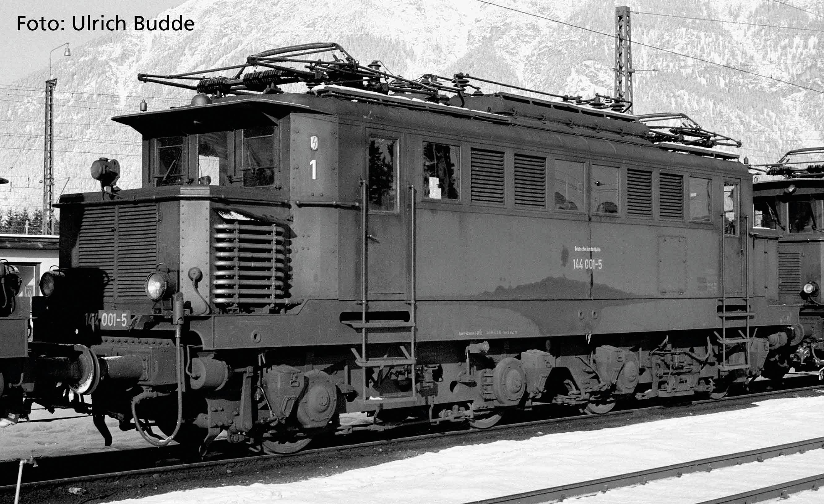 Eine historische Elektrolokomotive, seitlich auf Schienen abgebildet, mit eingeschneiten Bergen im Hintergrund. Foto von Ulrich Budde.