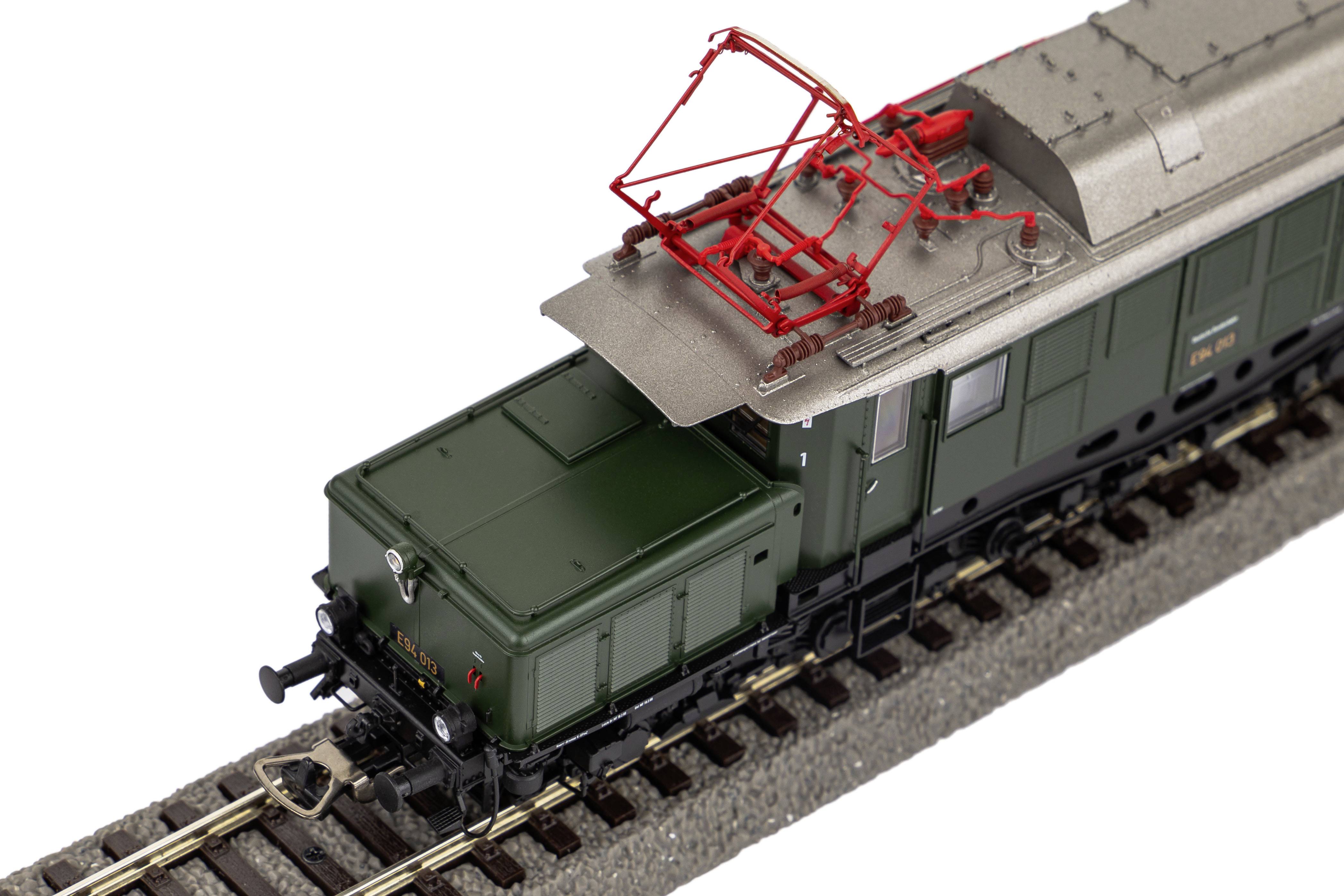 Ein Modellzuglokomotive mit einem roten Stromabnehmer auf einer Schiene. Die Lokomotive ist in Grün und Grau gehalten.