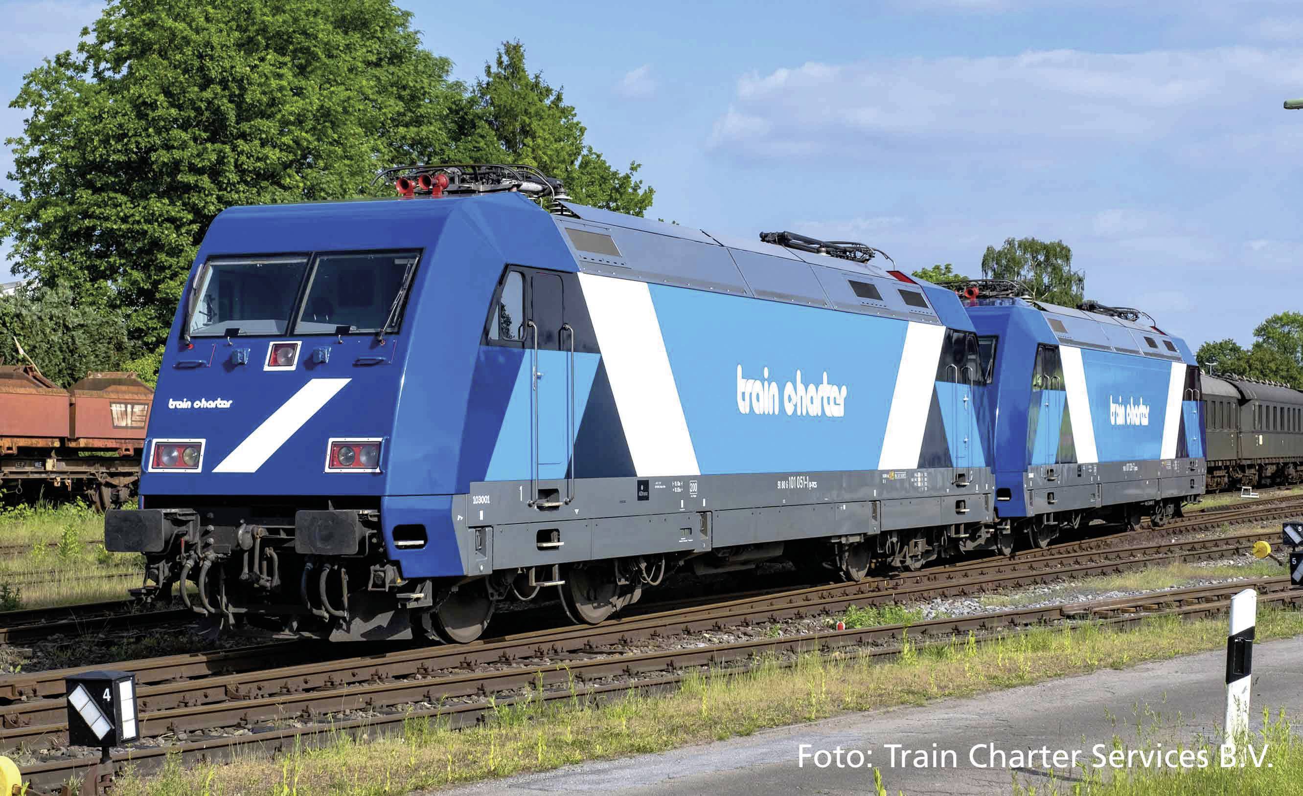 Zwei blaue, moderne Lokomotiven mit Aufschrift 'train charter' auf Eisenbahngleisen. Hintergrund zeigt Bäume und blauen Himmel.