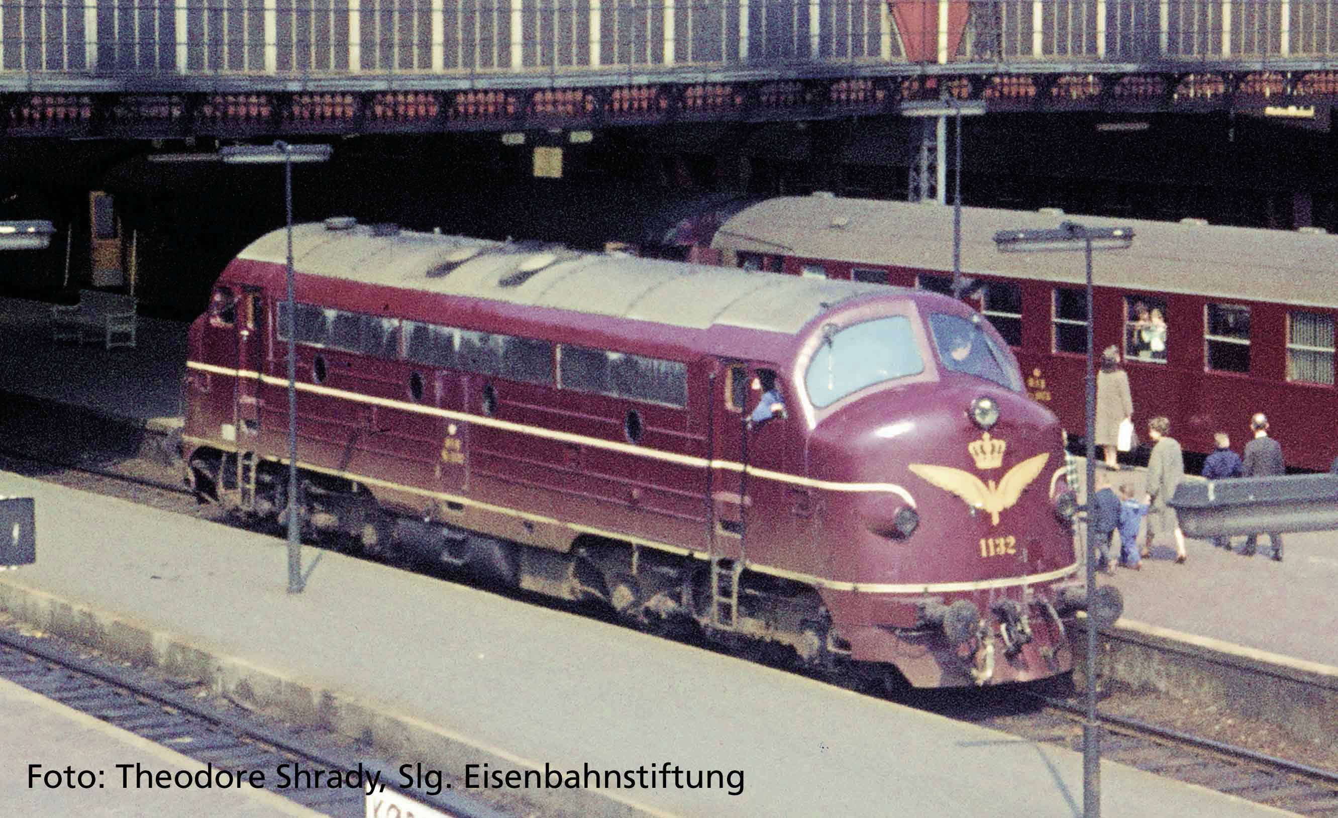 Eine rote Diesellokomotive steht auf einem Bahnsteig. Im Hintergrund sind Menschen zu sehen, die auf den Zug warten.