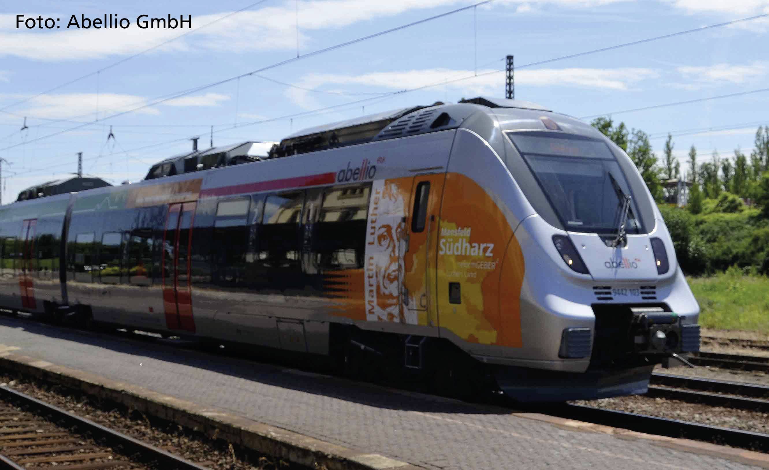 Ein moderner Regionalzug auf einem Bahngleis, im Hintergrund Strommasten und bewölkter Himmel. Zug hat bunte Werbegrafik 'Südharz'.