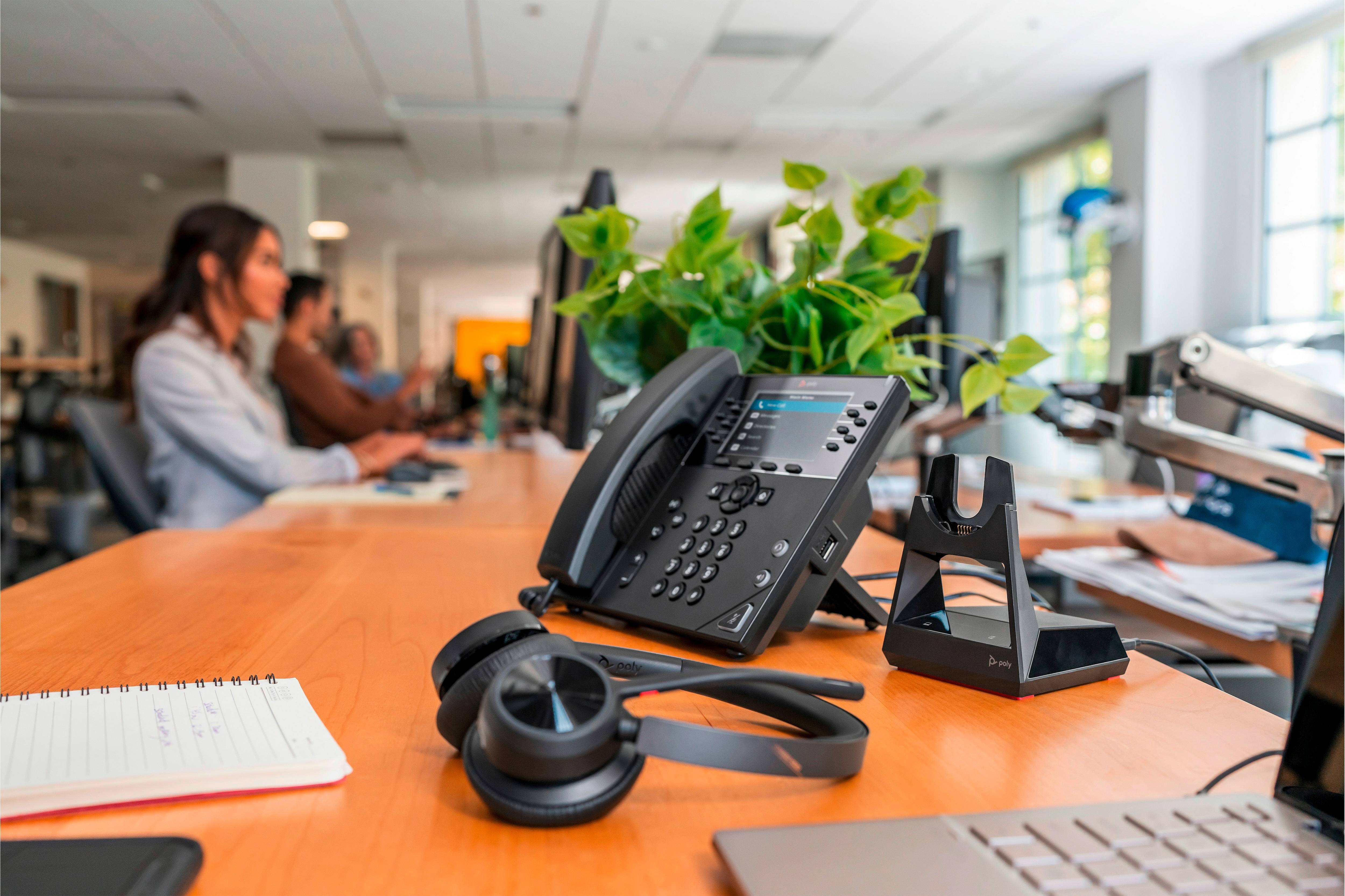 Ein Telefon und Kopfhörer liegen auf einem Schreibtisch in einem belebten Büro. Im Hintergrund arbeiten Personen an Computern.