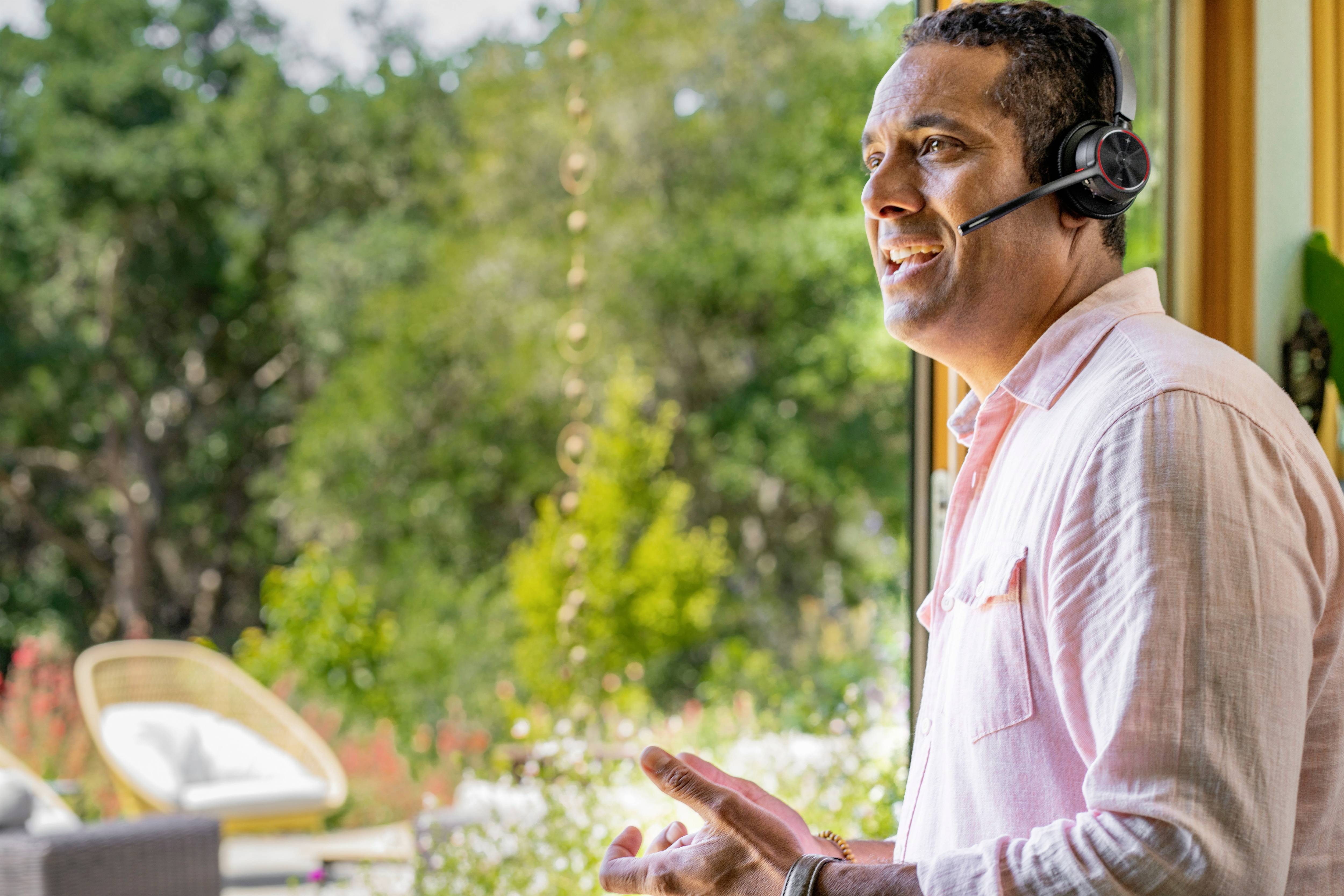 Ein Mann mit Headset telefoniert lächelnd in einem hellen Raum mit großen Fenstern. Draußen sind Bäume und ein Garten zu sehen.