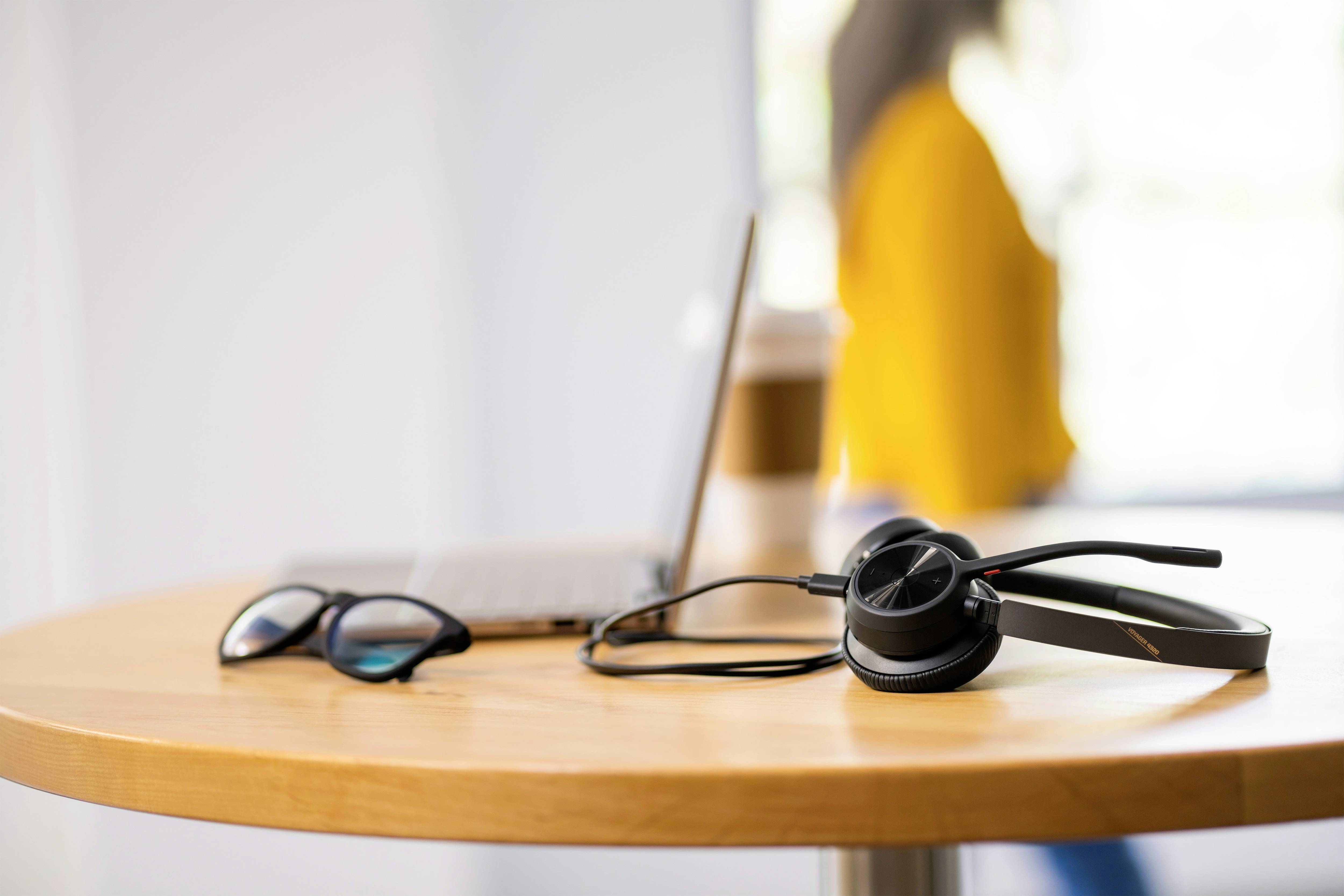 Ein runder Tisch mit Laptop, Brille und einem Headset. Eine verschwommene Person im Hintergrund. Fokus auf Arbeitsumgebung.