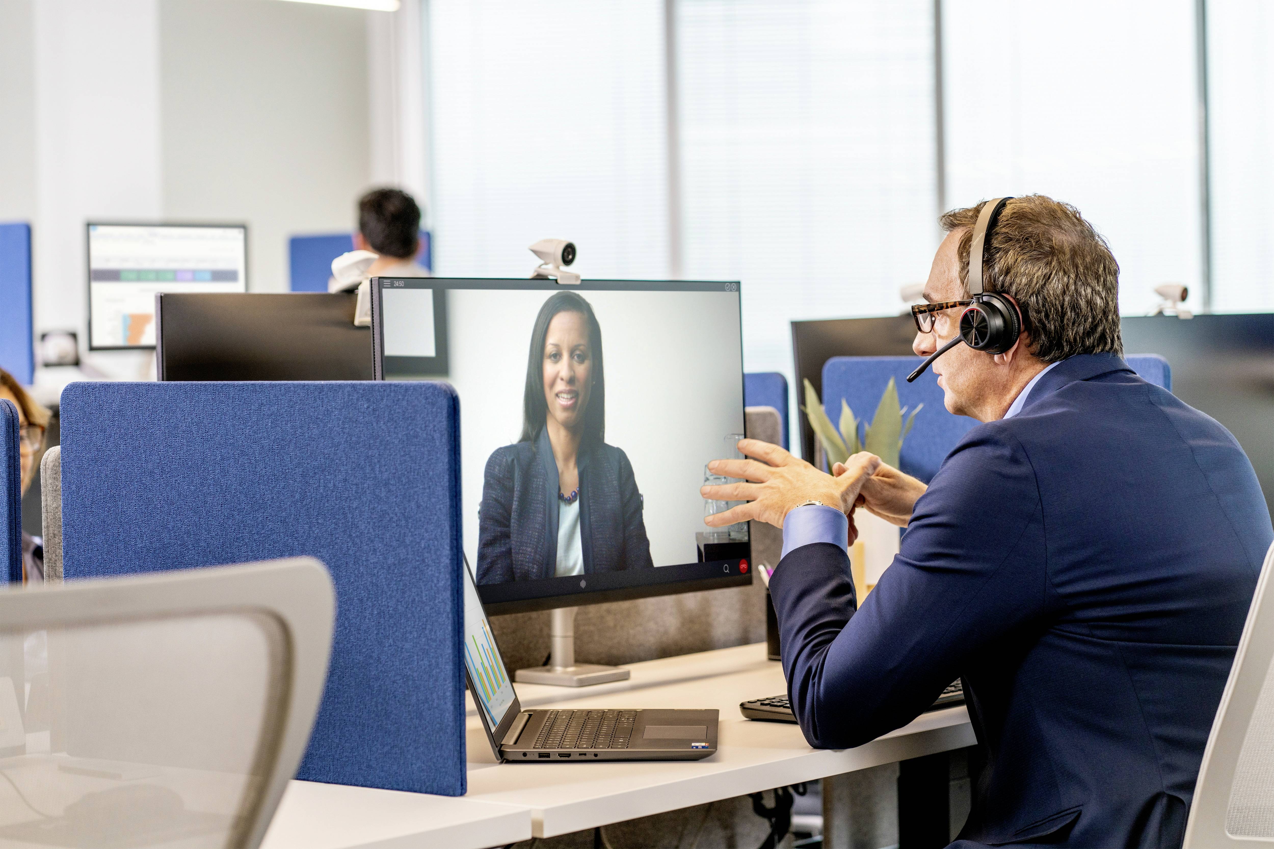 Ein Geschäftsmann mit Headset führt ein Videogespräch am Computer in einem modernen Büro. Im Hintergrund arbeiten weitere Personen.