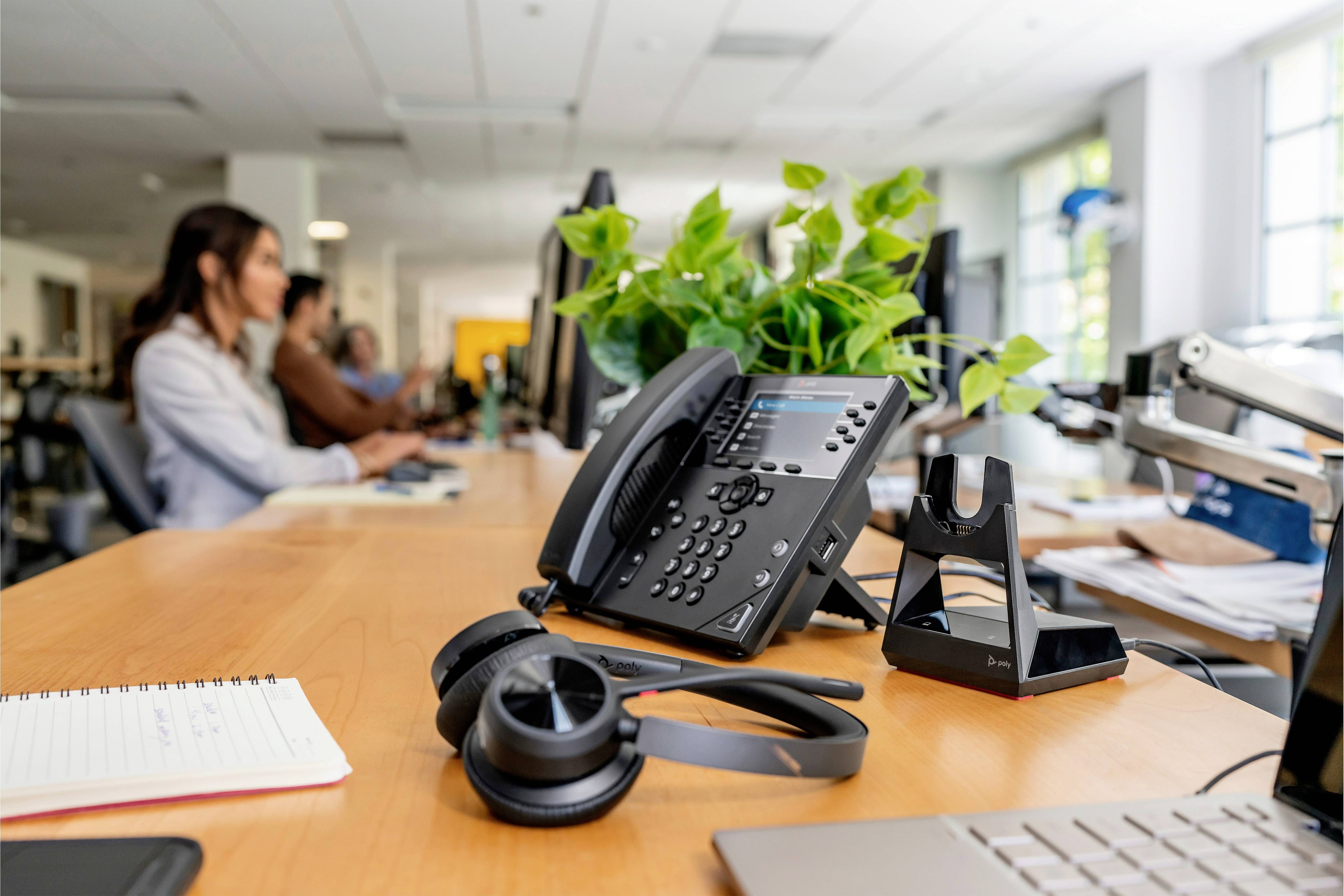 Ein Schreibtisch in einem Büro mit einem Telefon, Kopfhörern, einem Notizblock und Pflanzen im Vordergrund. Personen arbeiten im Hintergrund.