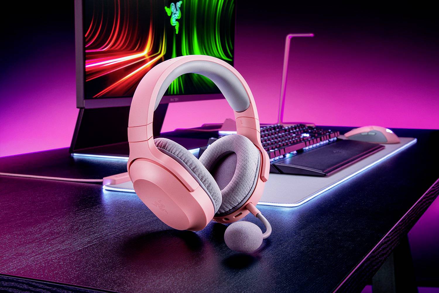 Ein rosa Gaming-Headset liegt auf einem beleuchteten Schreibtisch vor einem Monitor mit leuchtendem Bildschirmschoner und Tastatur.