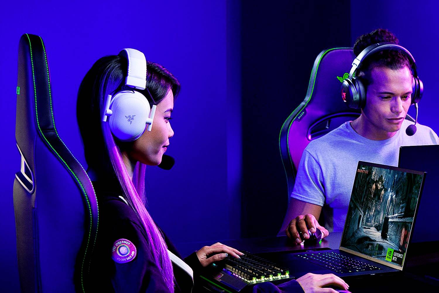 Zwei Personen spielen nebeneinander Videospiele an Computern mit Kopfhörern und Mikrofonen, in einem blau beleuchteten Raum.