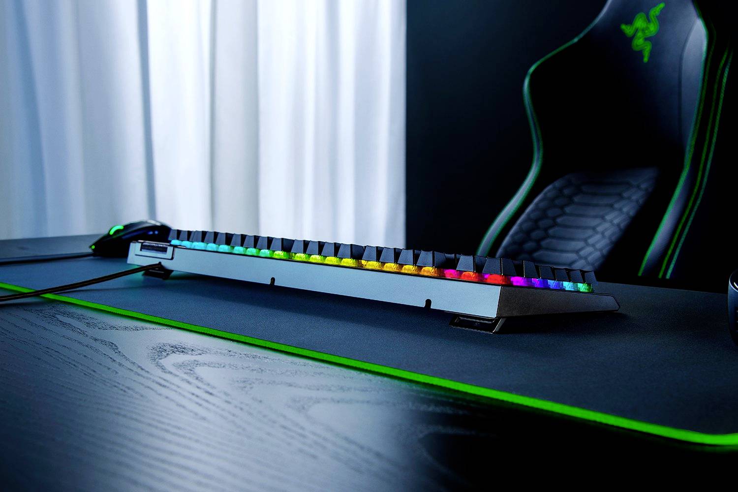 Eine Gaming-Tastatur mit farbiger RGB-Hintergrundbeleuchtung auf einem Schreibtisch neben einer Gaming-Maus und einem Gaming-Stuhl.