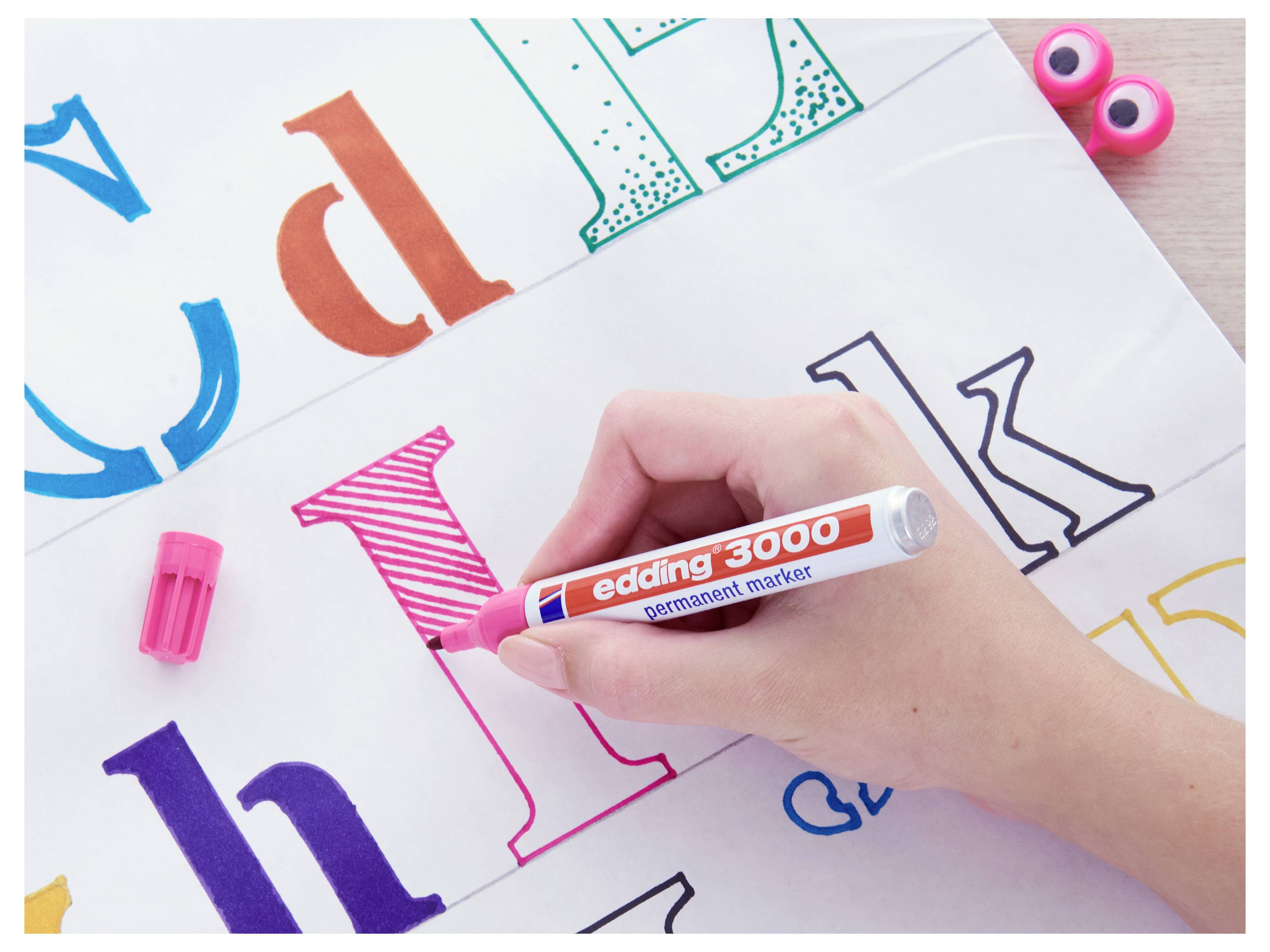 Hand, die einen rosa Marker hält und einen großen Kleinbuchstaben i auf Alphabet-Übungsblättern färbt, während andere farbige Buchstaben sichtbar sind