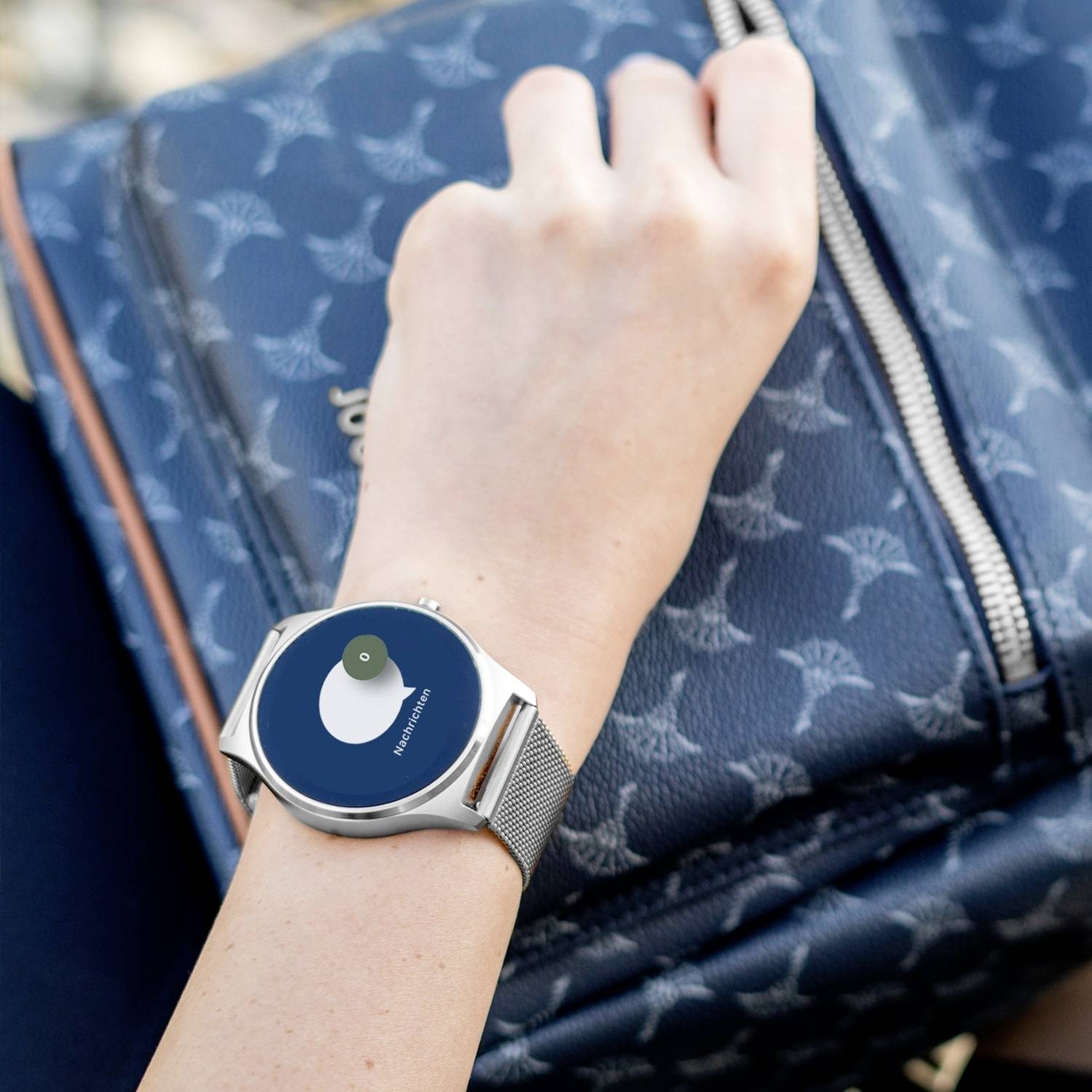 Eine Hand trägt eine Smartwatch mit einem blauen Zifferblatt, das eine Benachrichtigung zeigt. Der Hintergrund ist teilweise ein Rucksack.