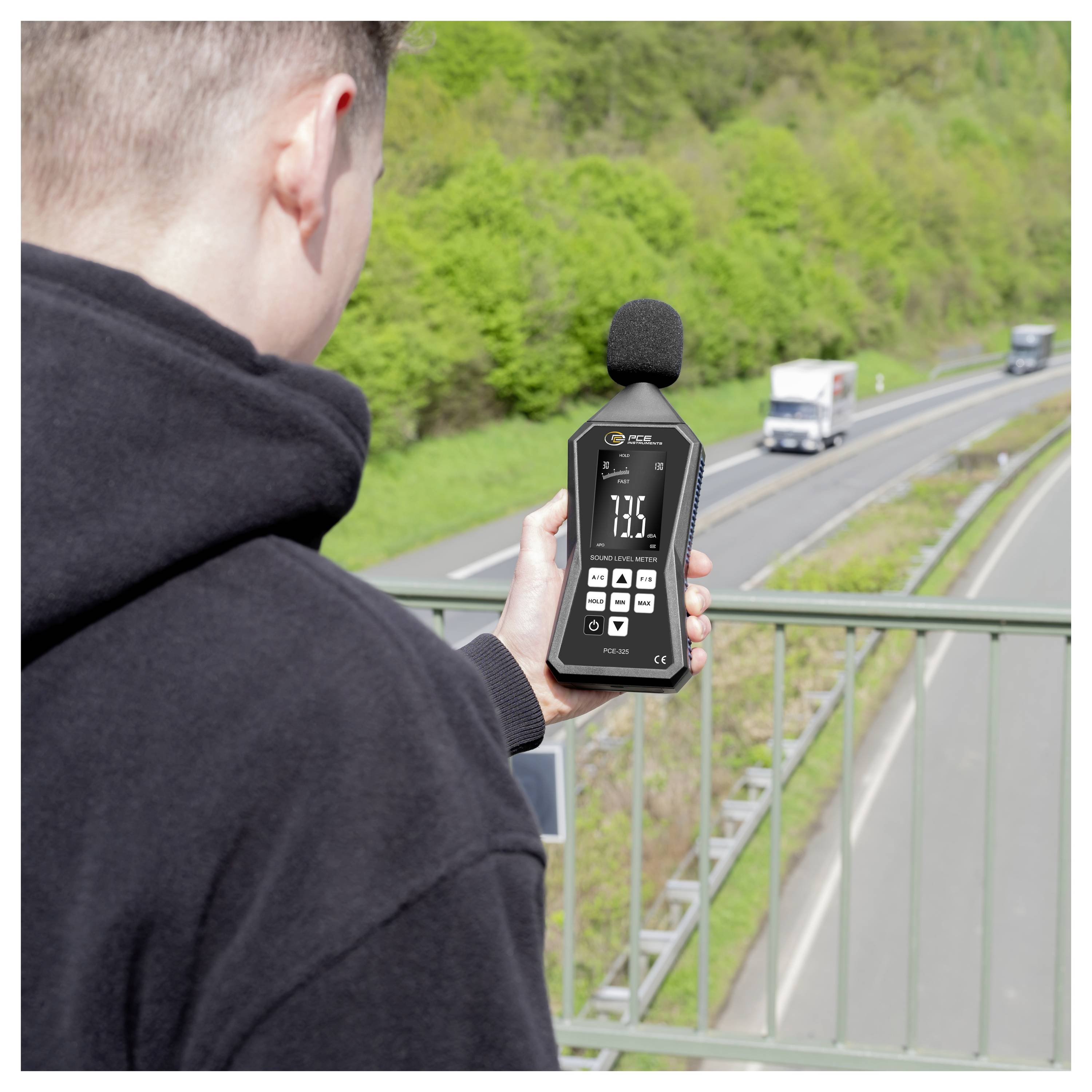 Person mit einem Schallpegelmesser zur Messung von Verkehrslärm neben einer Autobahn, mit vorbeifahrenden Lastkraftwagen im Hintergrund.