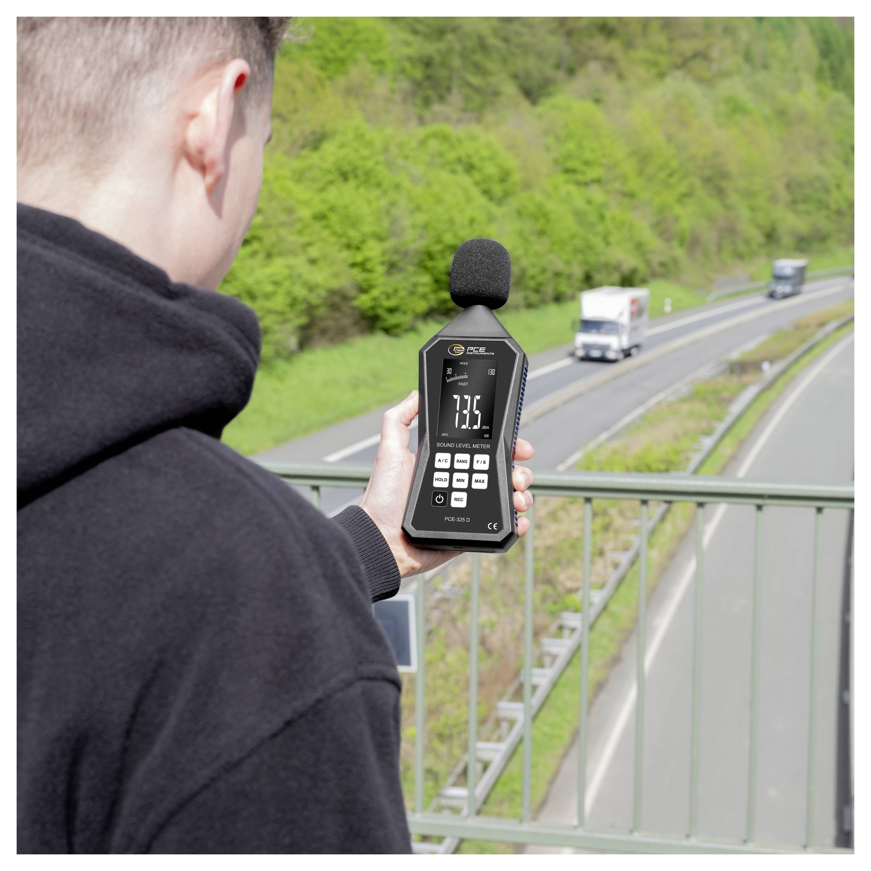 Person misst Straßenlärm mit einem tragbaren Schallpegelmessgerät neben einer Autobahn, wobei ein Messwert von 73,5 dB angezeigt wird.
