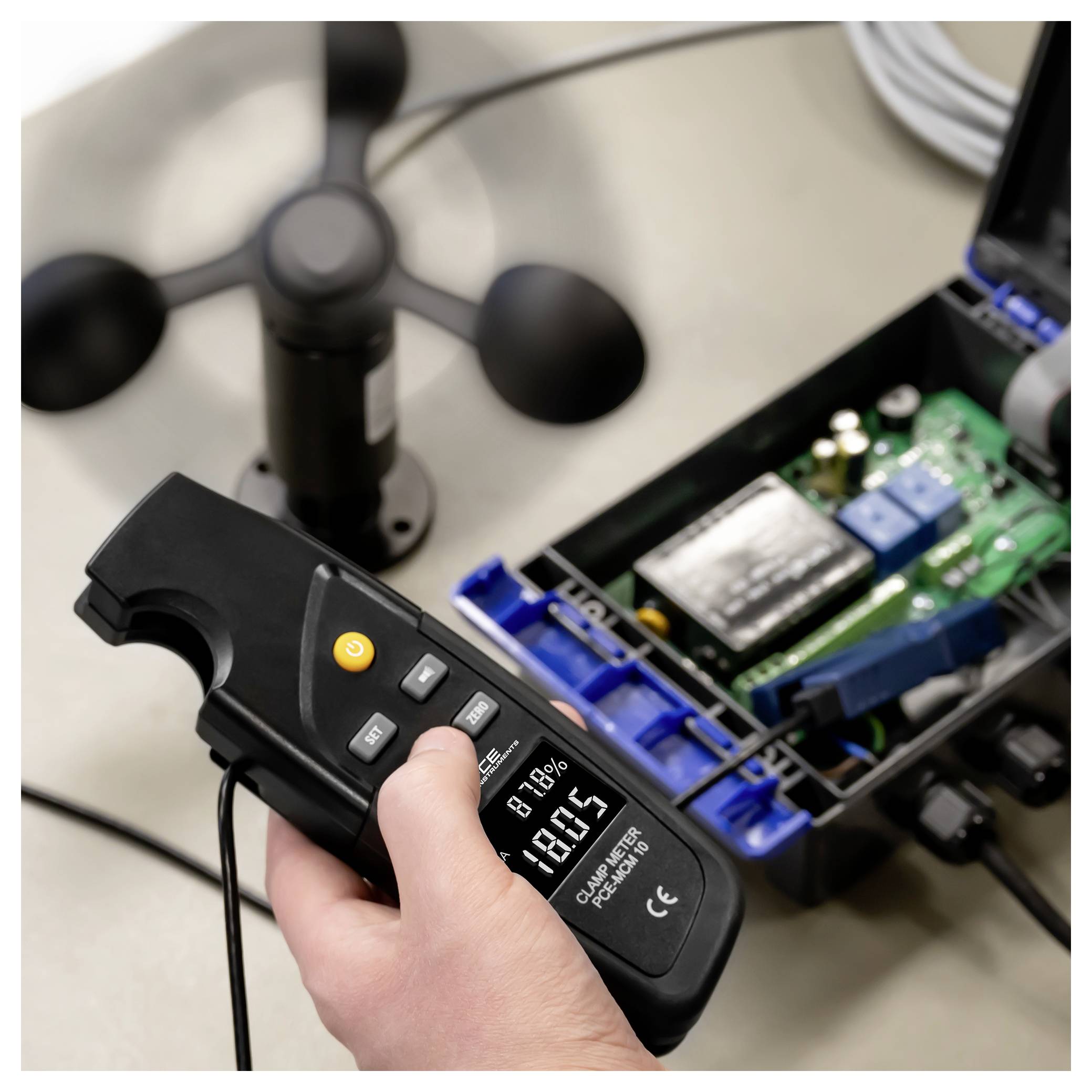 Eine Hand, die ein digitales Messgerät hält, das die Spannung an einem offenen elektronischen Gerät mit einer Leiterplatte und einem Lüfter im Hintergrund anzeigt.