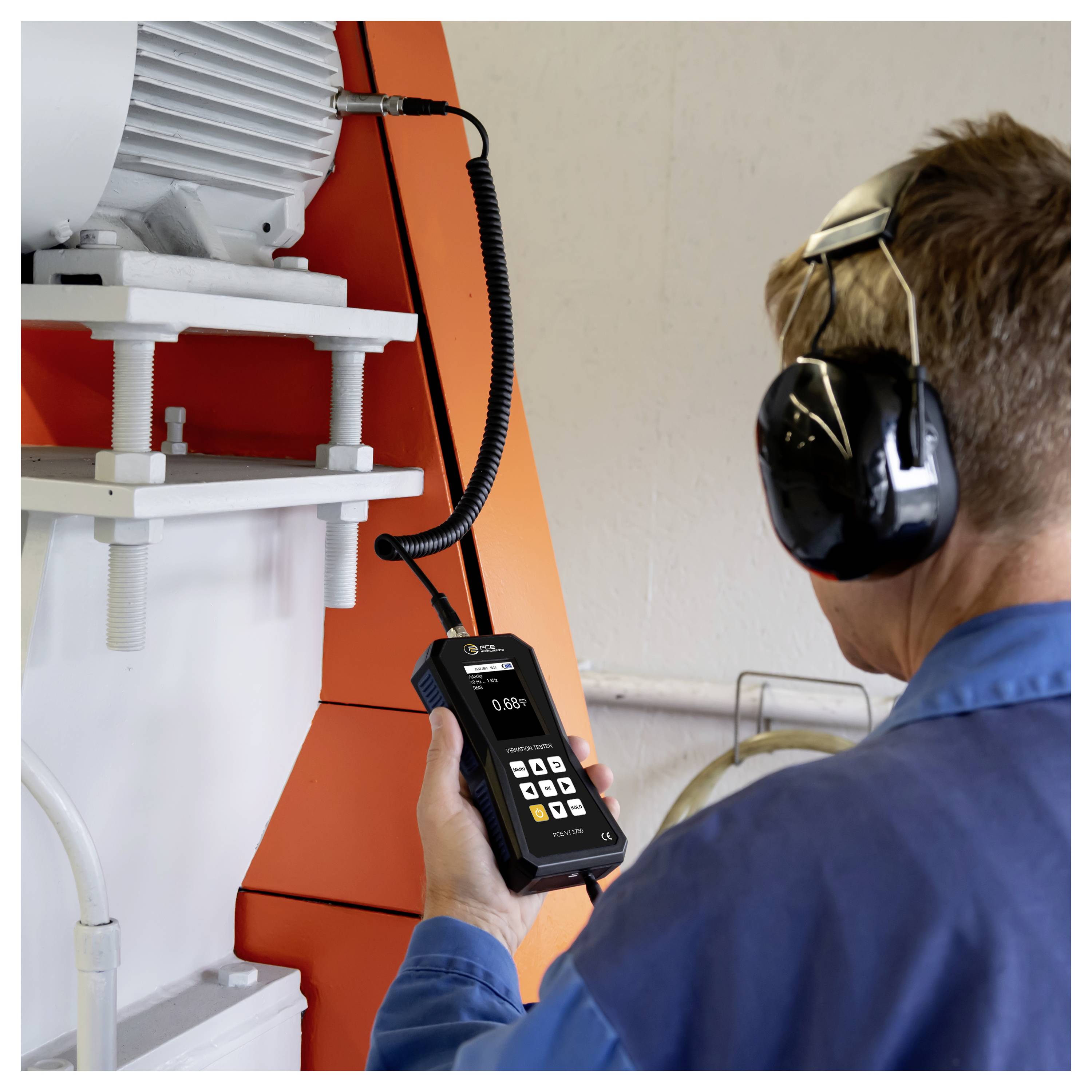 Techniker mit Gehörschutz nutzt ein tragbares Messgerät zur Überprüfung von Industriemaschinen neben einem großen Motor und einem orangefarbenen Bedienfeld.