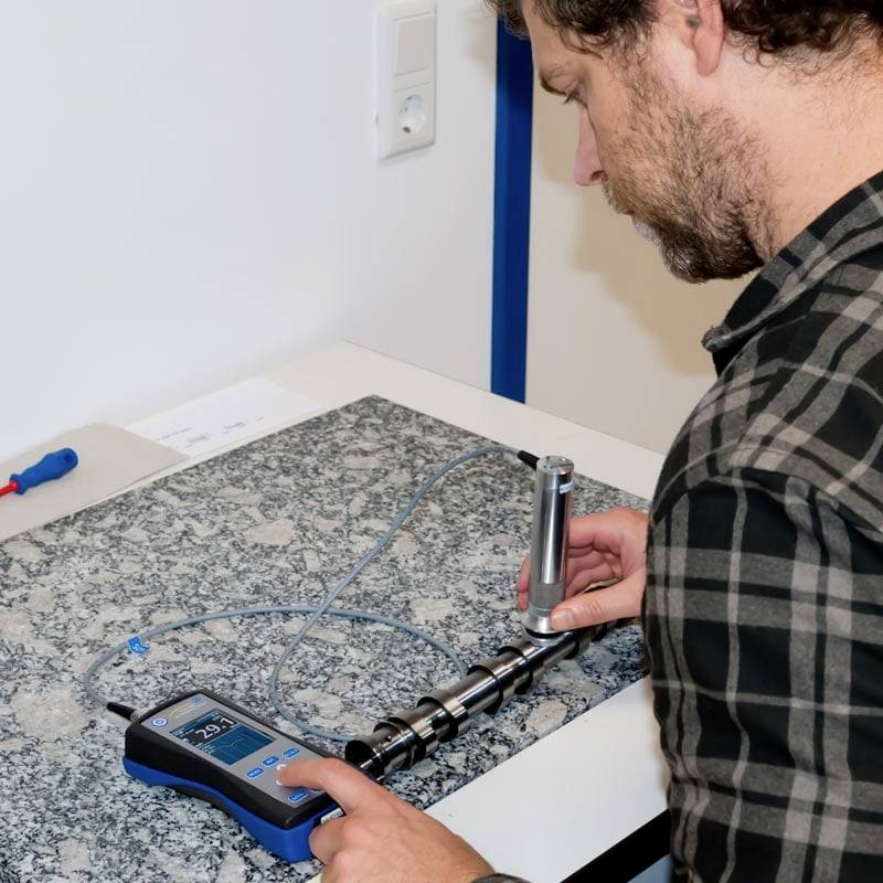 Ein Mann misst mit einem digitalen Messgerät die Spannung auf einem metallischen Schaltstab auf einem Tisch.