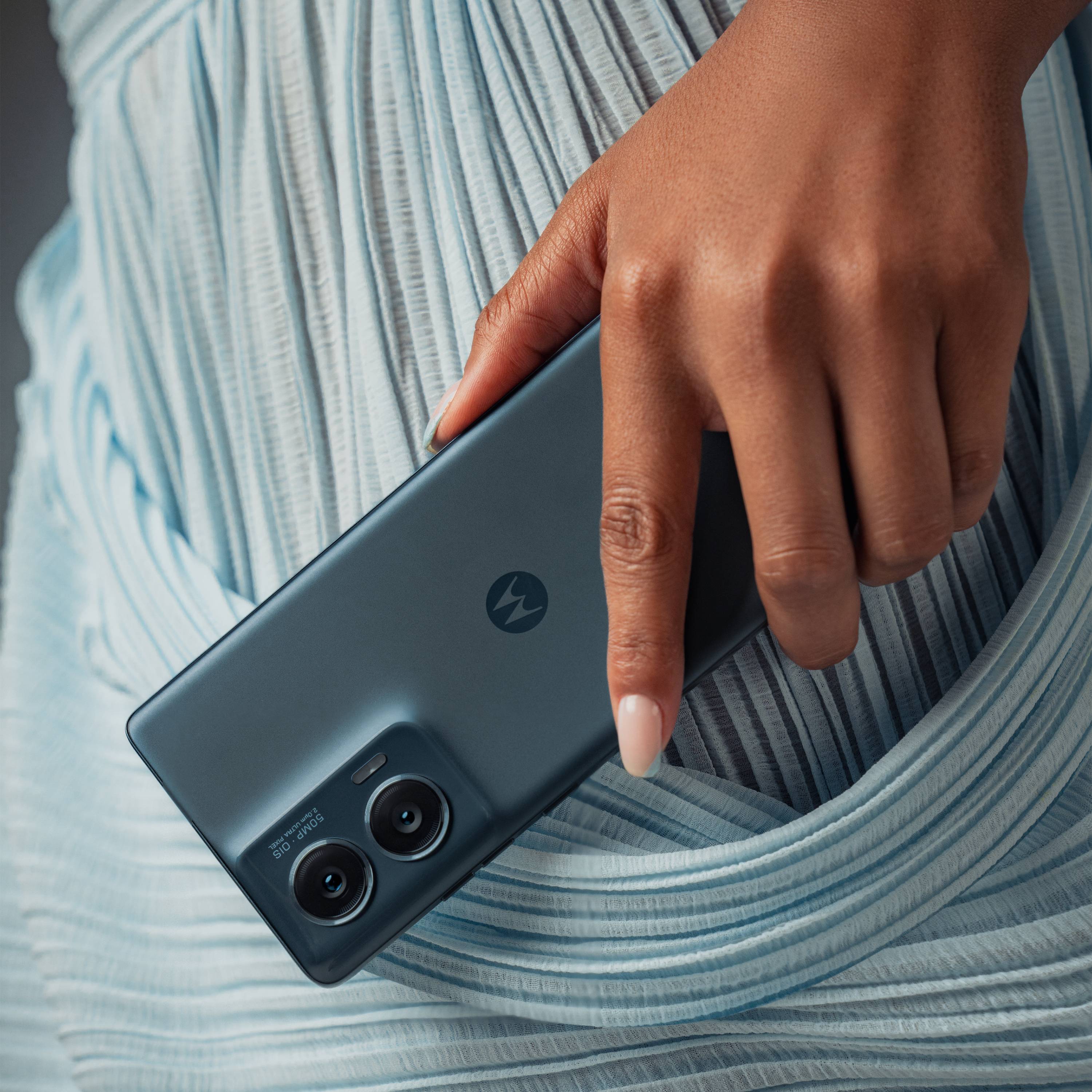 Eine Hand hält ein modernes Smartphone mit zwei Kameralinsen auf der Rückseite. Die Person trägt ein hellblaues Kleid.