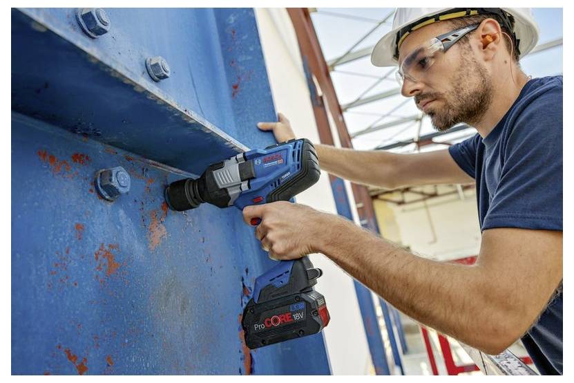 Bauarbeiter benutzt einen kabellosen Bohrer, um einen großen blauen Stahlträger auf einer Baustelle zu befestigen, trägt einen Schutzhelm und eine Schutzbrille.