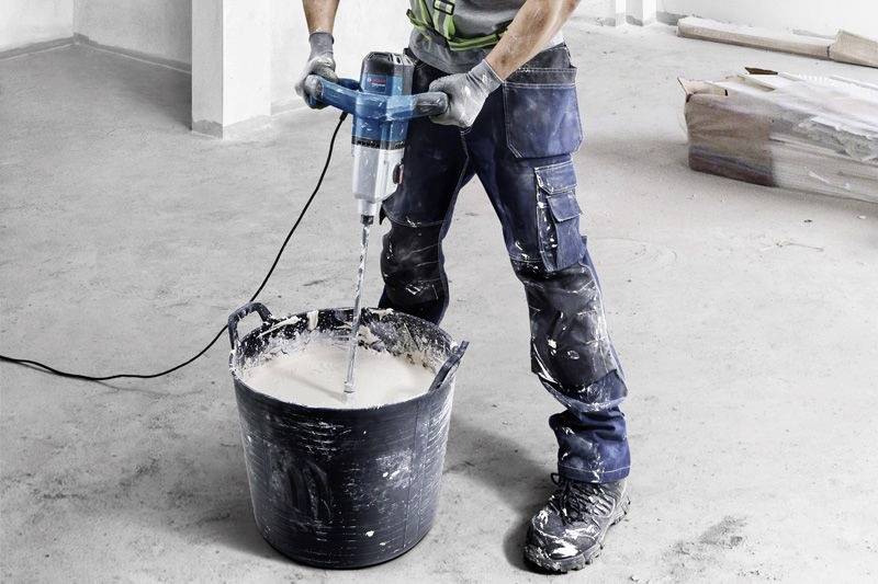 Eine Person mischt Baustoffe in einem Eimer mit einem elektrischen Rührgerät. Der Boden ist sichtbar mit Bauwerkzeugen und Materialien im Hintergrund.