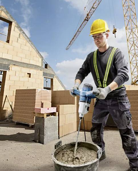 Ein Bauarbeiter mit Schutzhelm und Sicherheitsweste mischt Zement mit einem elektrischen Rührwerk auf einer Baustelle mit Kränen im Hintergrund.