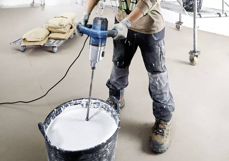 Eine Person mischt mit einem elektrischen Rührgerät weiße Farbe in einem großen Eimer. Im Hintergrund liegen Säcke auf einer Plattform.