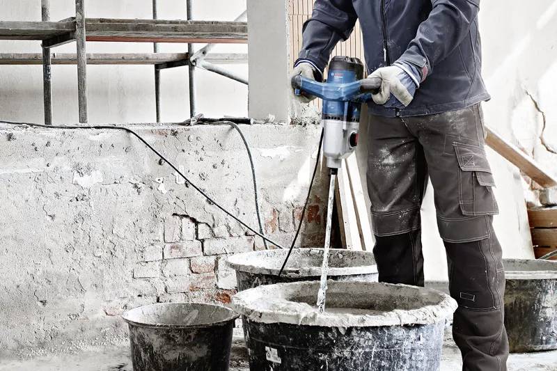 Ein Bauarbeiter mischt mit einem elektrischen Rührgerät Zement in einem Eimer. Im Hintergrund sind Wände und ein Gerüst sichtbar.