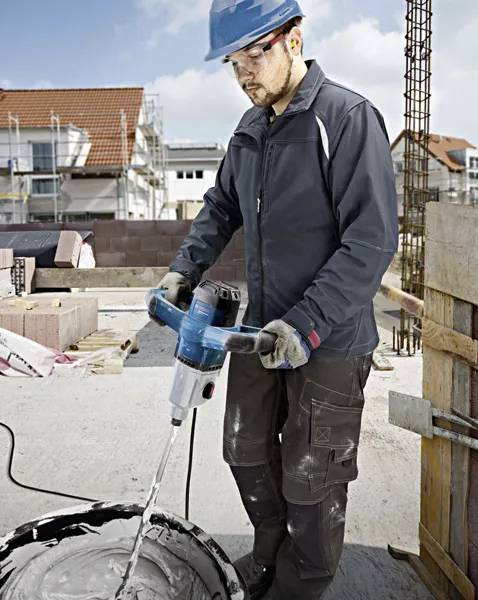 Ein Bauarbeiter mischt Zement auf einer Baustelle mit einem elektrischen Handmischer. Im Hintergrund sind Baugerüste und Ziegelstapel.