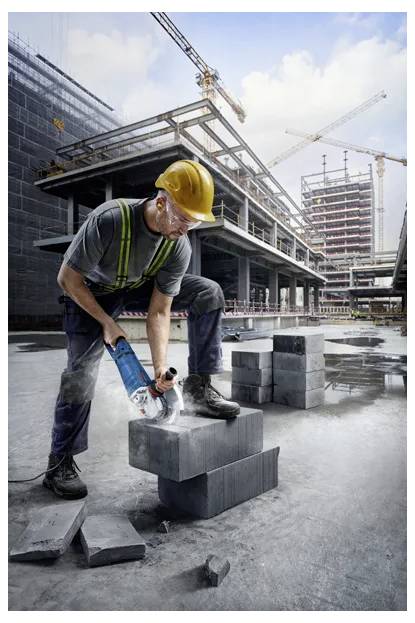 Ein Bauarbeiter mit gelbem Helm und Schutzausrüstung schneidet Betonblöcke mit einem Elektrowerkzeug auf einer großen Baustelle mit Kränen.