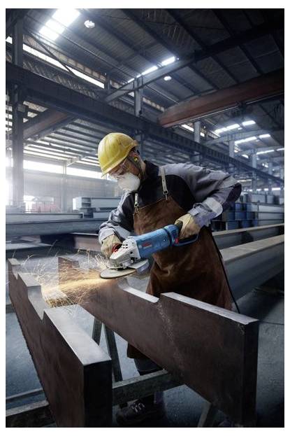 Ein Fabrikarbeiter in Schutzausrüstung schleift einen Metallträger mit einem Elektrowerkzeug und erzeugt Funken in einer geräumigen, industriellen Lagerhalle.