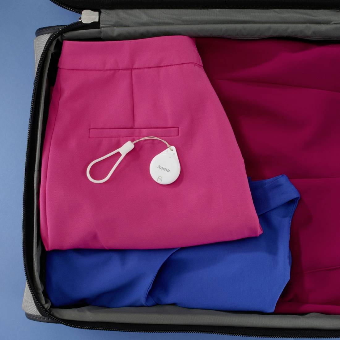 Ein offener Koffer zeigt eine rosa Hose mit einem weißen Tracker, daneben ein blaues Kleidungsstück.