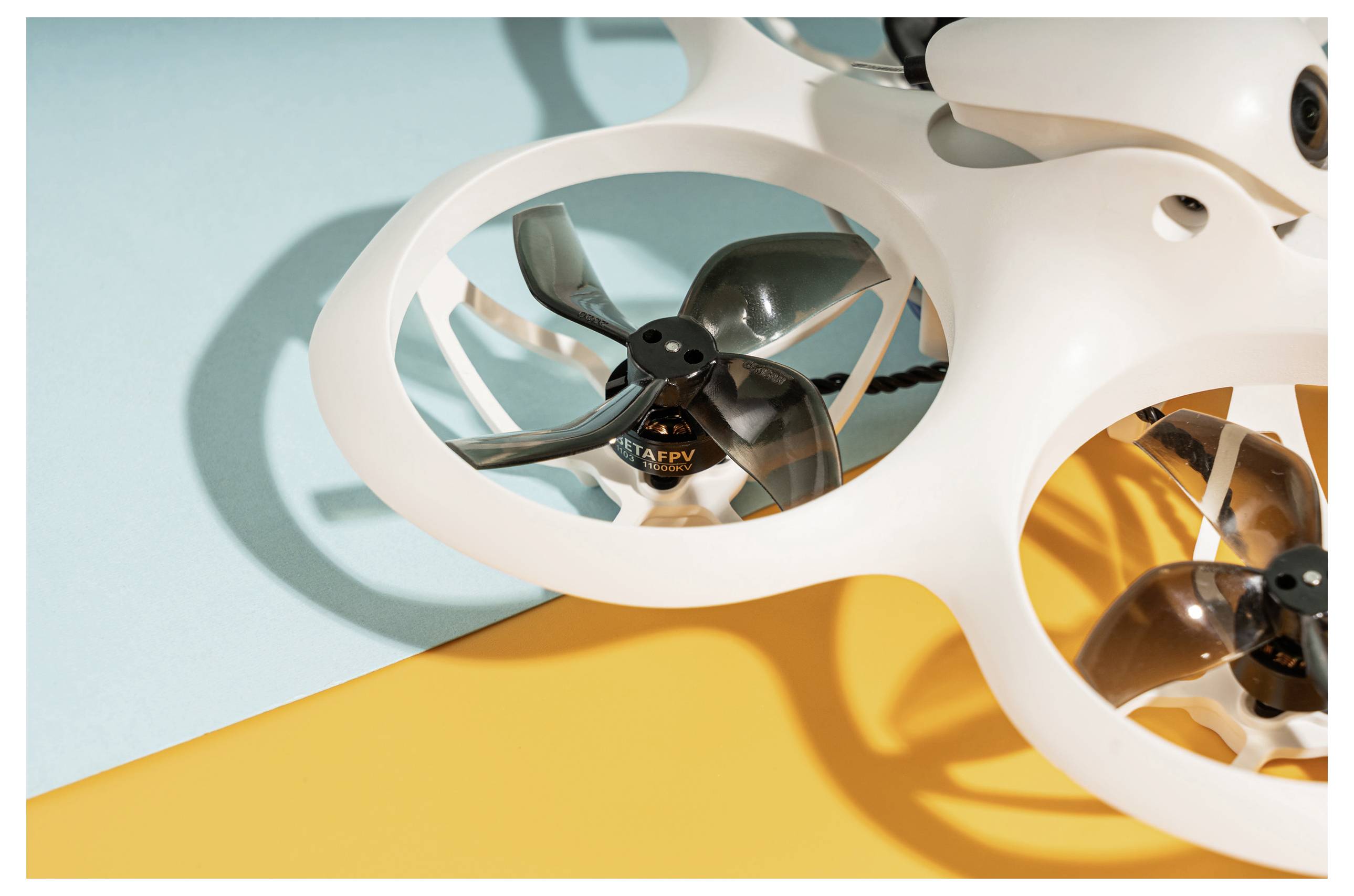 Eine kleine Drohne mit schwarzen Propellern in einem weißen Rahmen sitzt auf einer blau-orangefarbenen Oberfläche und wirft kontrastierende Schatten.