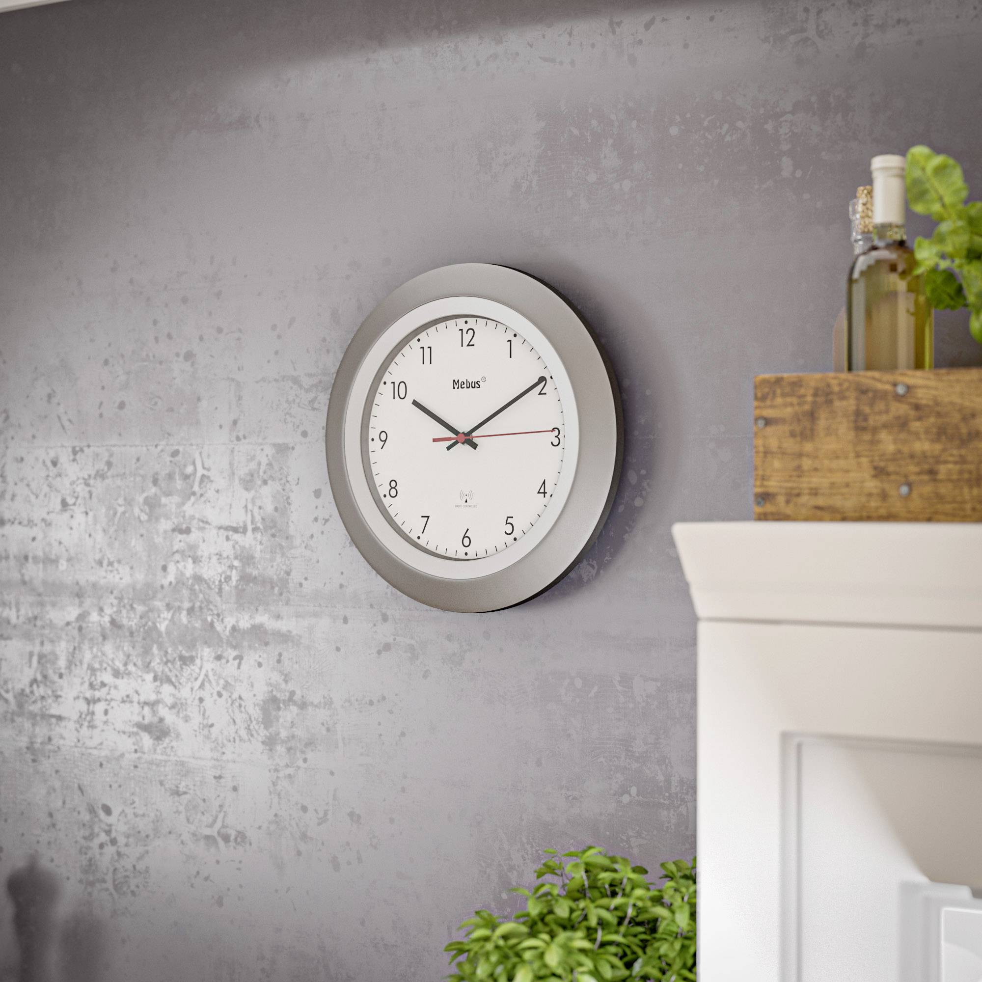 Eine runde Wanduhr mit weißen Ziffernblättern und schwarzen Zeigern hängt an einer grauen Wand. Links unten ist eine Pflanze zu sehen.
