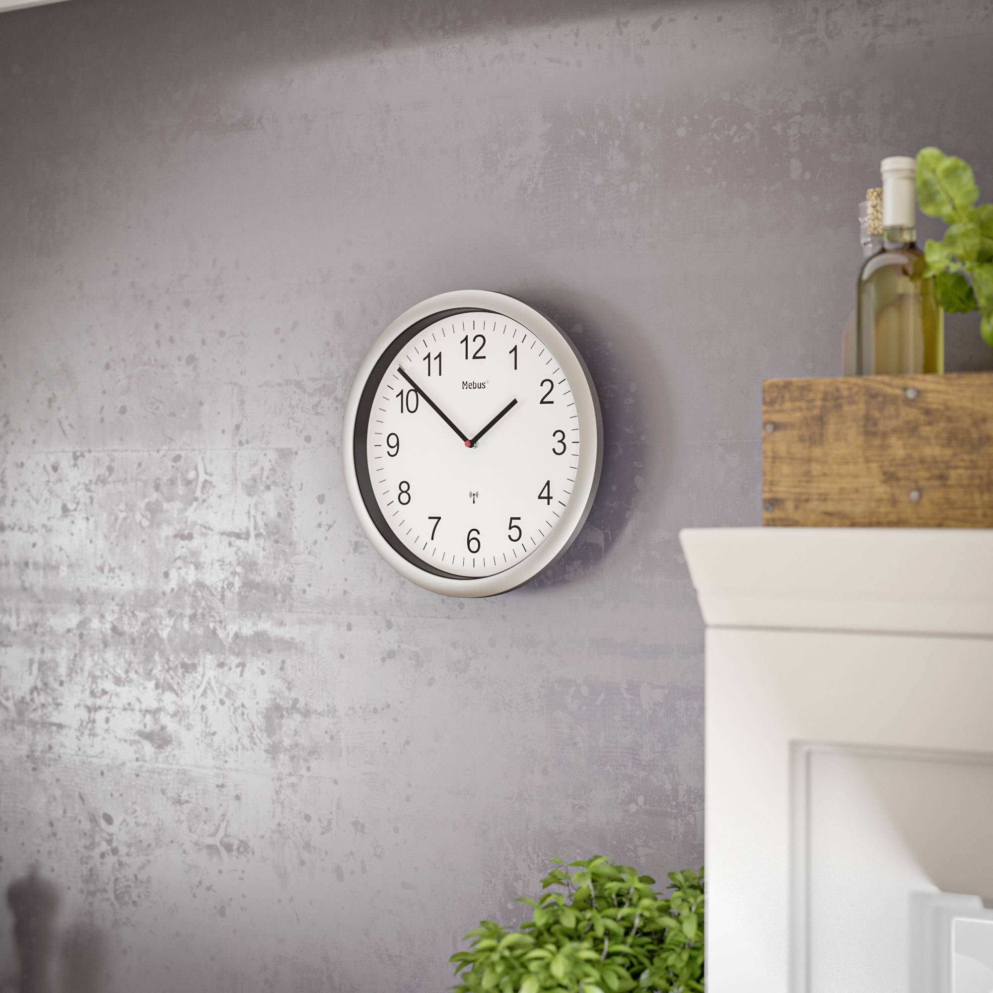 Eine runde Wanduhr mit schwarzem Rahmen zeigt 10:10 Uhr an. Sie hängt an einer grauen Wand neben einem Regal mit Pflanzen.