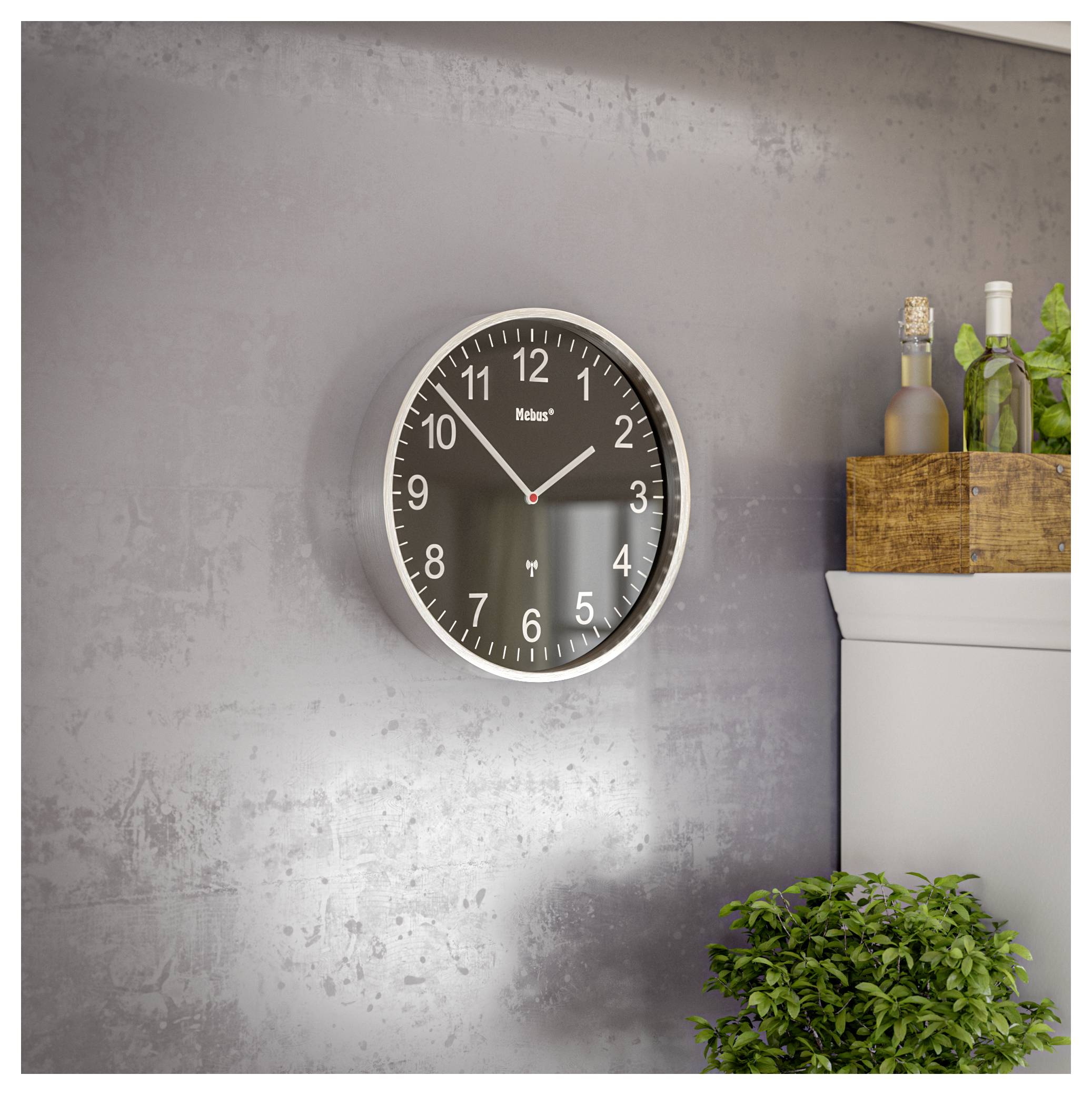 Eine runde Wanduhr mit weißen Ziffern und Zeigern auf dunklem Zifferblatt, die 10:10 Uhr anzeigt. Sie hängt an einer grauen Wand über einem Schrank mit Flaschen und Grünpflanzen.