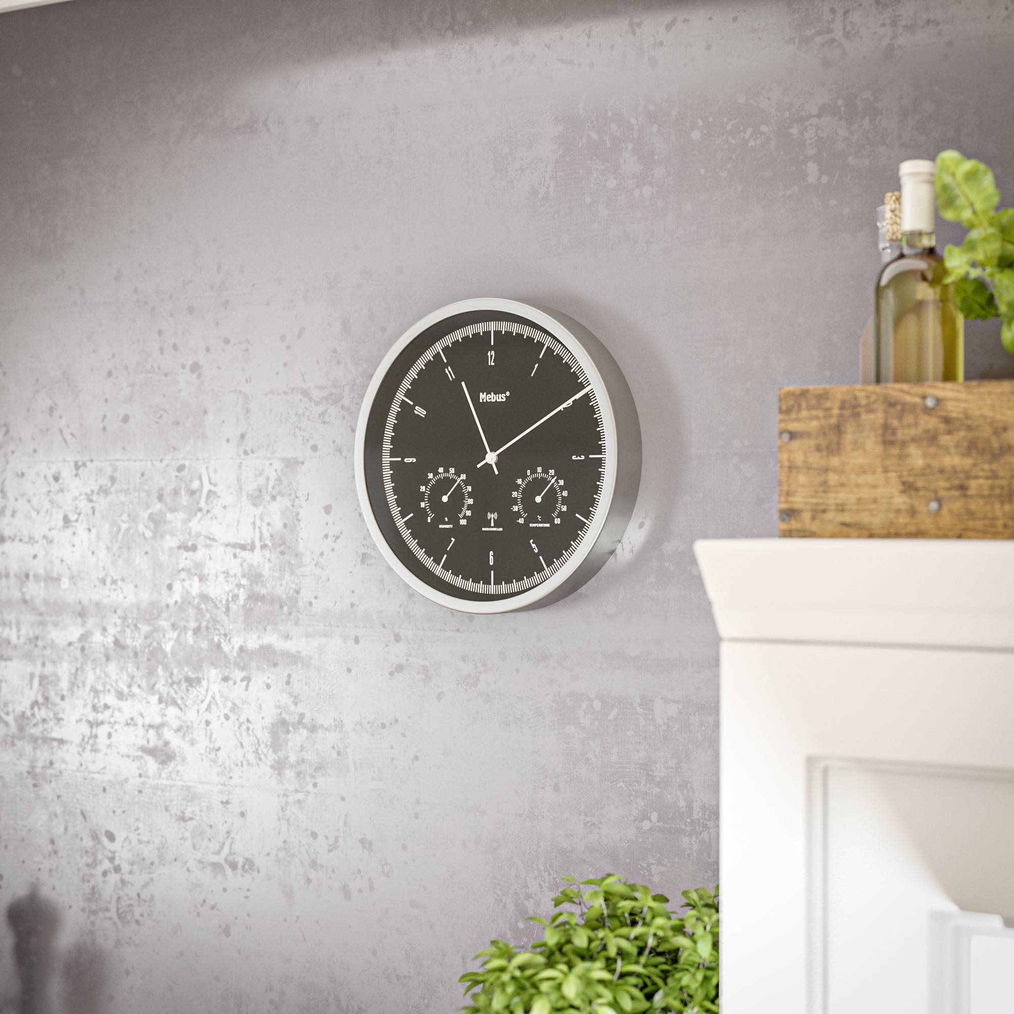 Eine schwarze Wanduhr mit weißen Zeigern und Ziffern hängt an einer grauen Wand. Daneben steht eine grüne Pflanze auf einem Schrank.