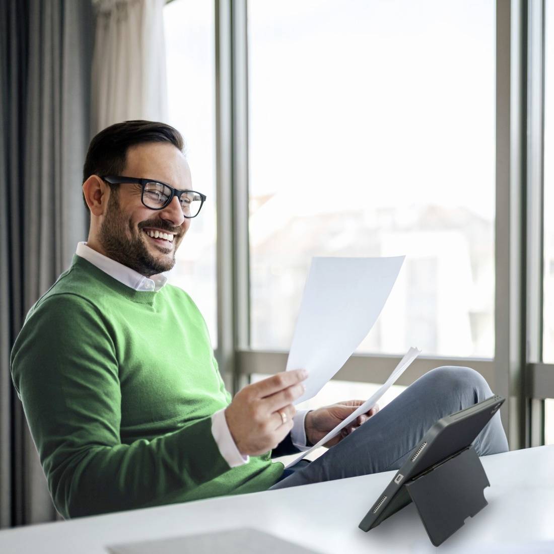 Ein Mann mit Brille und grünem Pullover sitzt entspannt lächelnd auf einem Sofa, hält ein Dokument und verwendet ein Tablet.