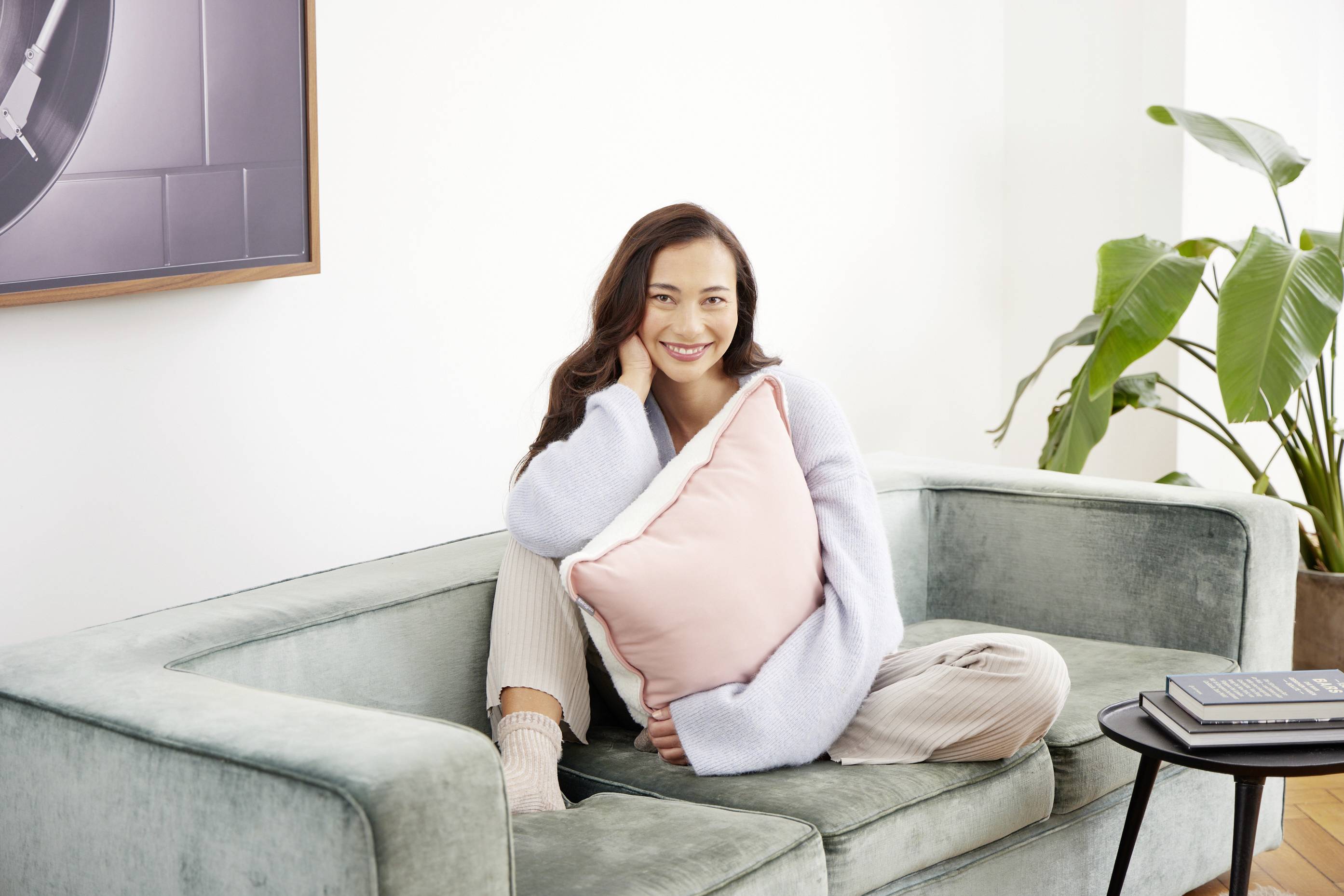 Eine lächelnde Person sitzt auf einem grünen Sofa, hält ein rosa Kissen. Im Hintergrund befinden sich eine Pflanze und ein gerahmtes Bild.