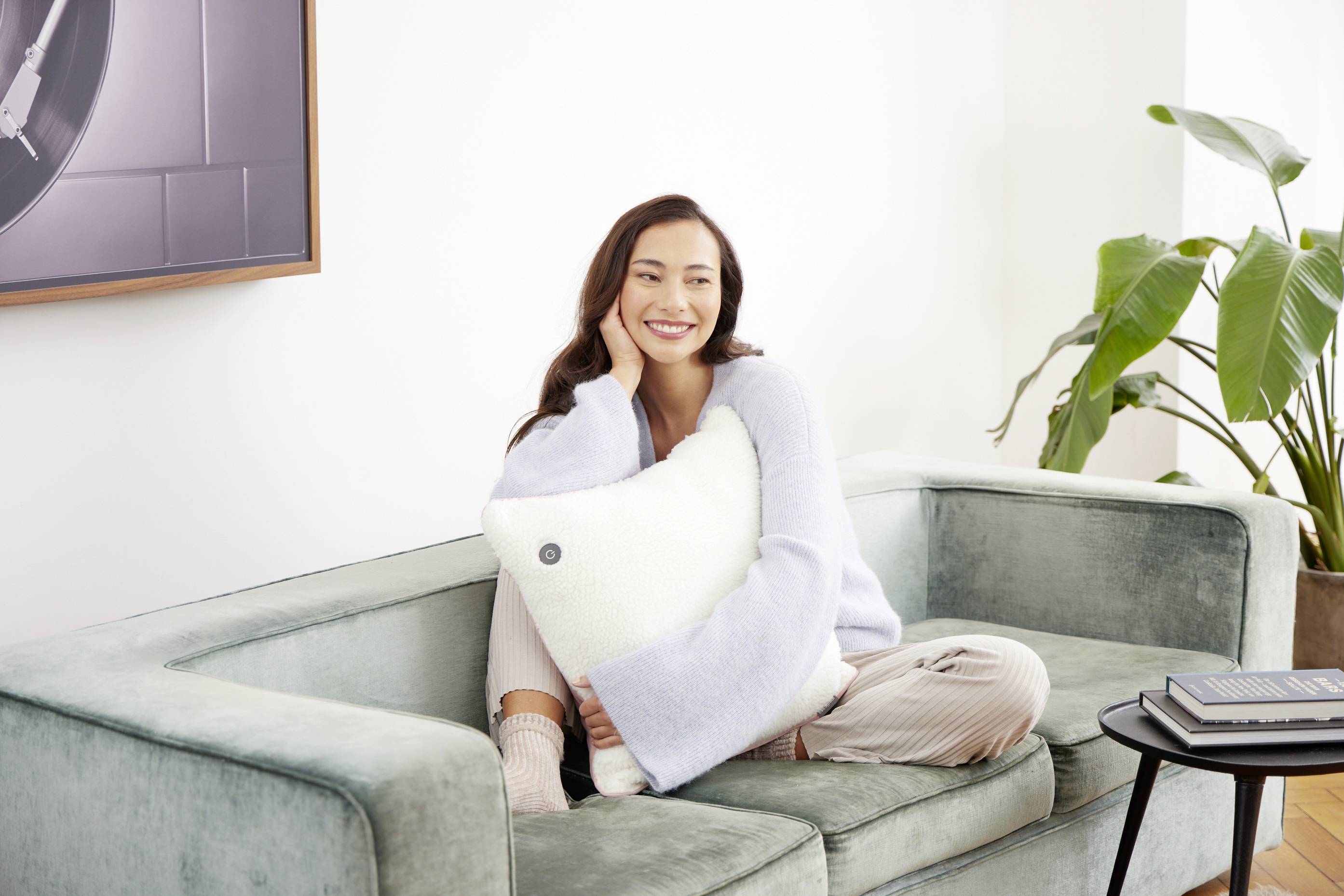 Frau sitzt lächelnd auf einem Sofa, hält ein Kissen und schaut entspannt aus. Im Hintergrund eine Pflanze und ein gerahmtes Bild.