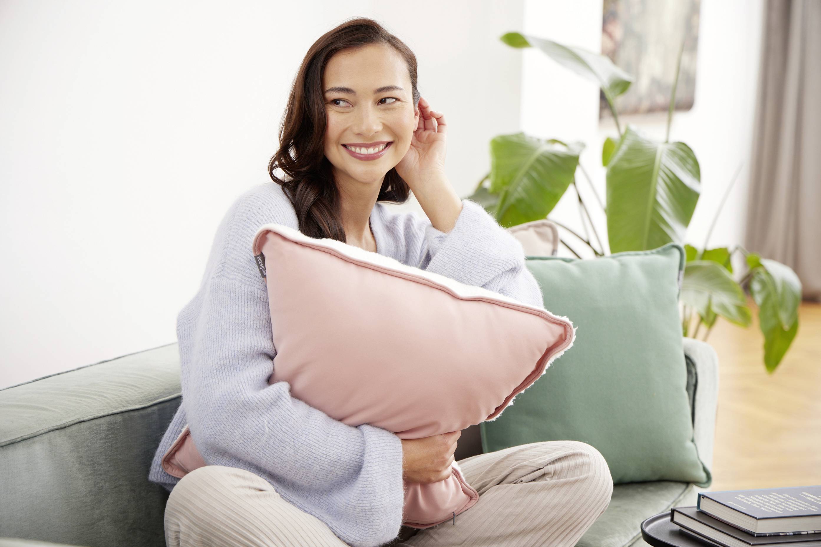 Eine lächelnde Frau sitzt auf einem Sofa und hält ein rosafarbenes Kissen. Im Hintergrund sind Pflanzen und ein Gemälde sichtbar.