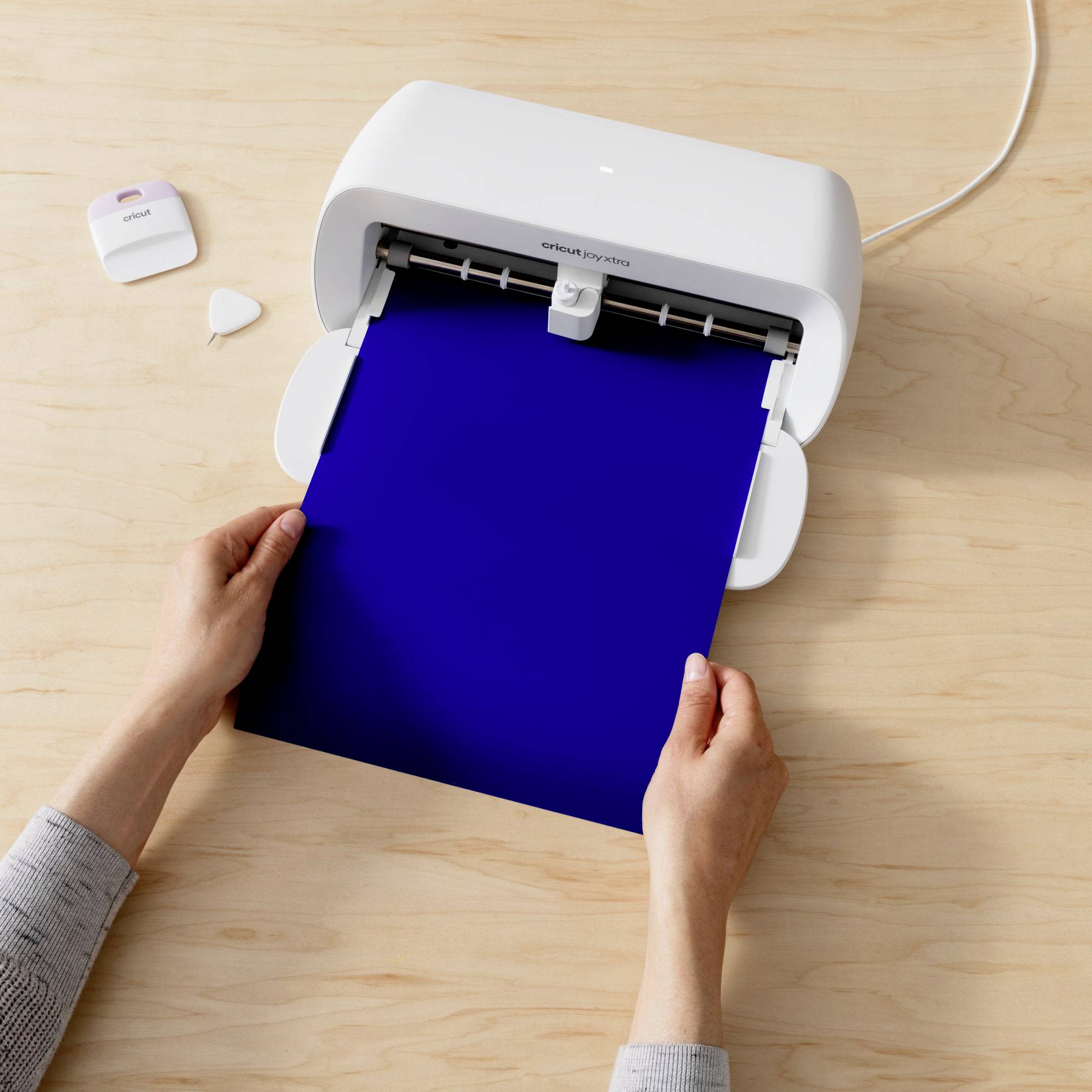 Eine Person legt ein blaues Blatt Papier in einen weißen Schneideplotter auf einem Tisch. Zubehör liegt daneben.